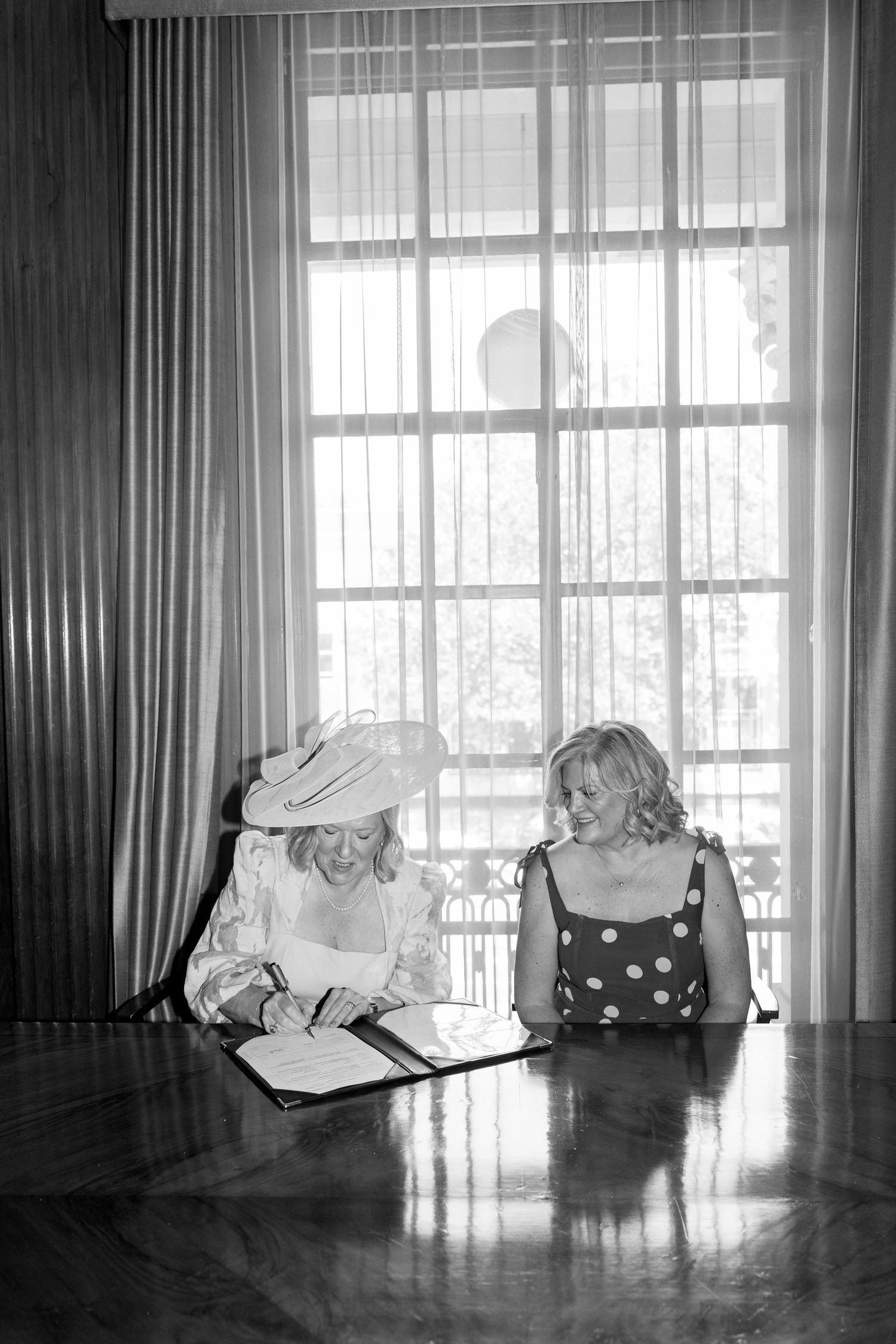 Two women smile while signing wedding documents at a table in front of a bright window.