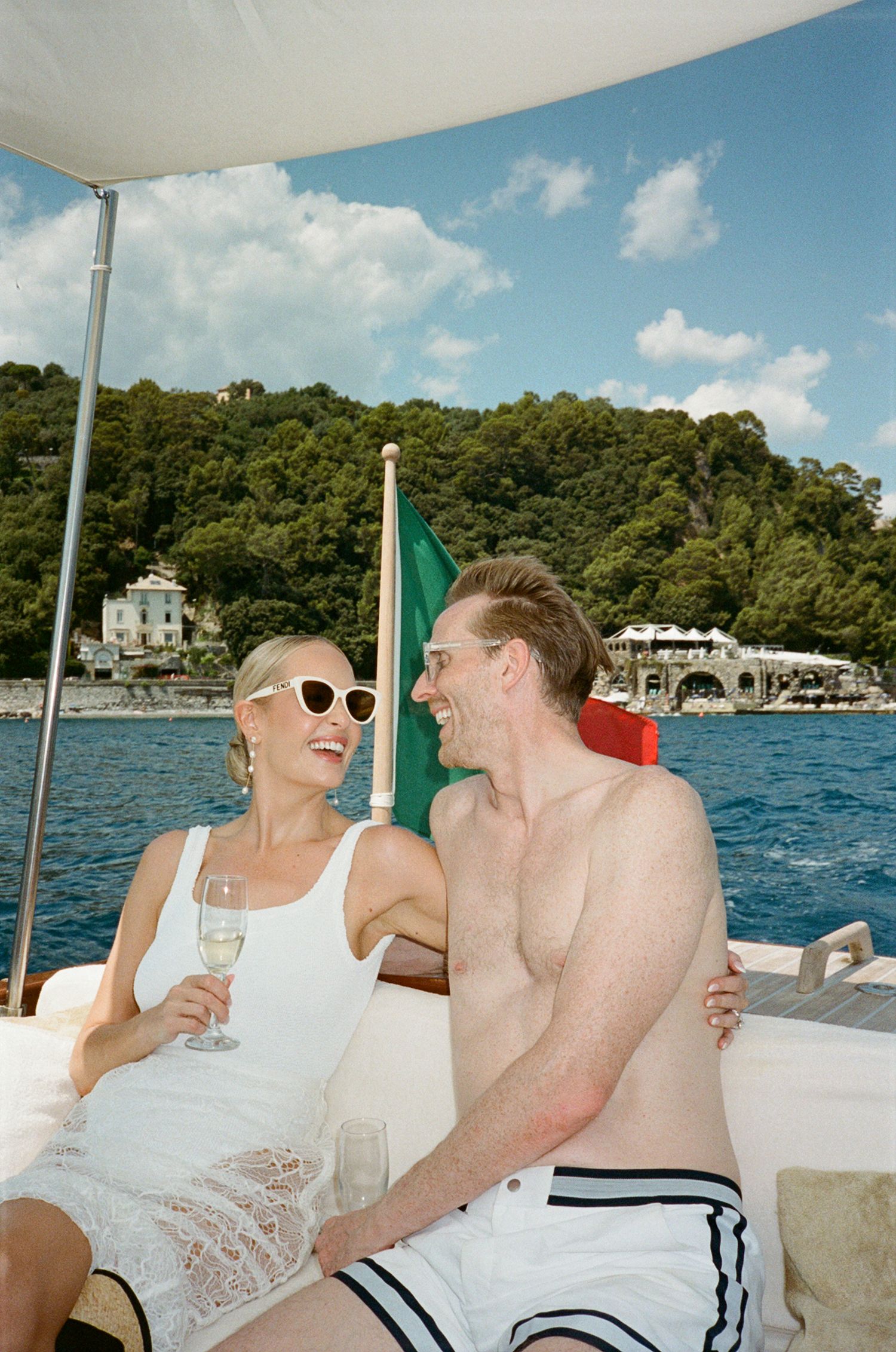 Couple enjoying champagne on a luxury boat in Portofino, Italy, with lush green hills and a villa in the background.