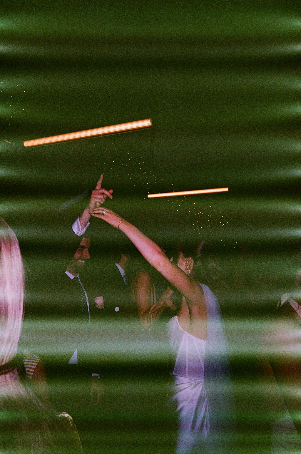 Bride in white dress raises glowing light bar on green-lit dance floor during wedding reception celebration.