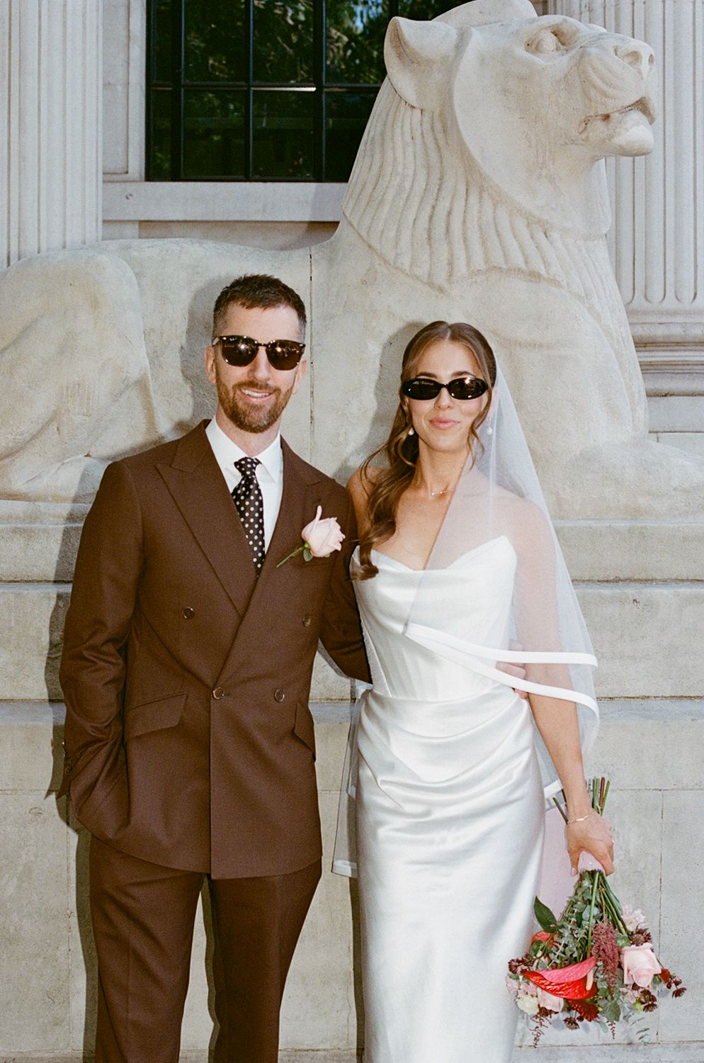 Stylish bride and groom in sunglasses pose before stone lion sculpture at elegant wedding venue.