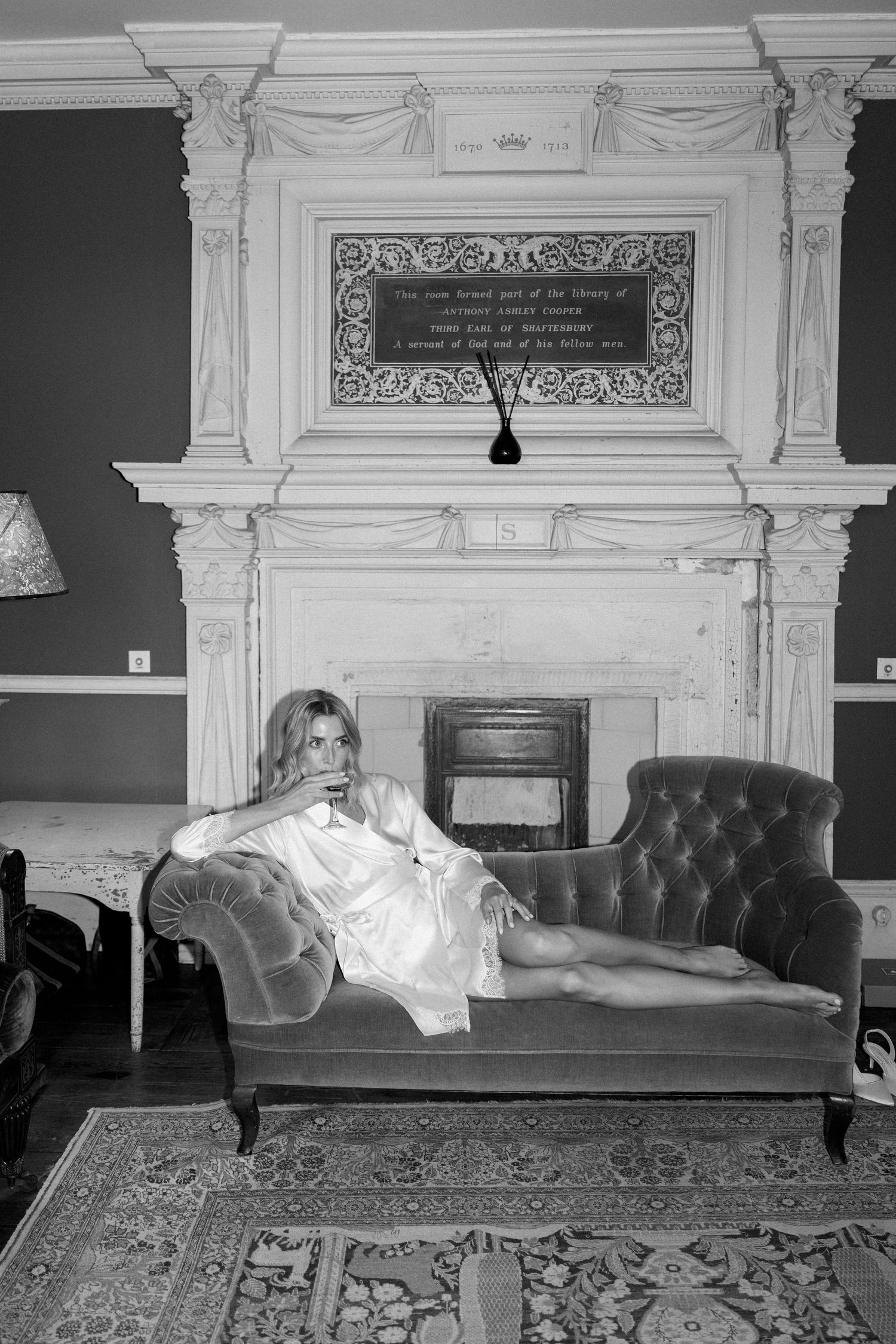 Woman in white silk robe reclining on tufted sofa before ornate fireplace in elegant manor.