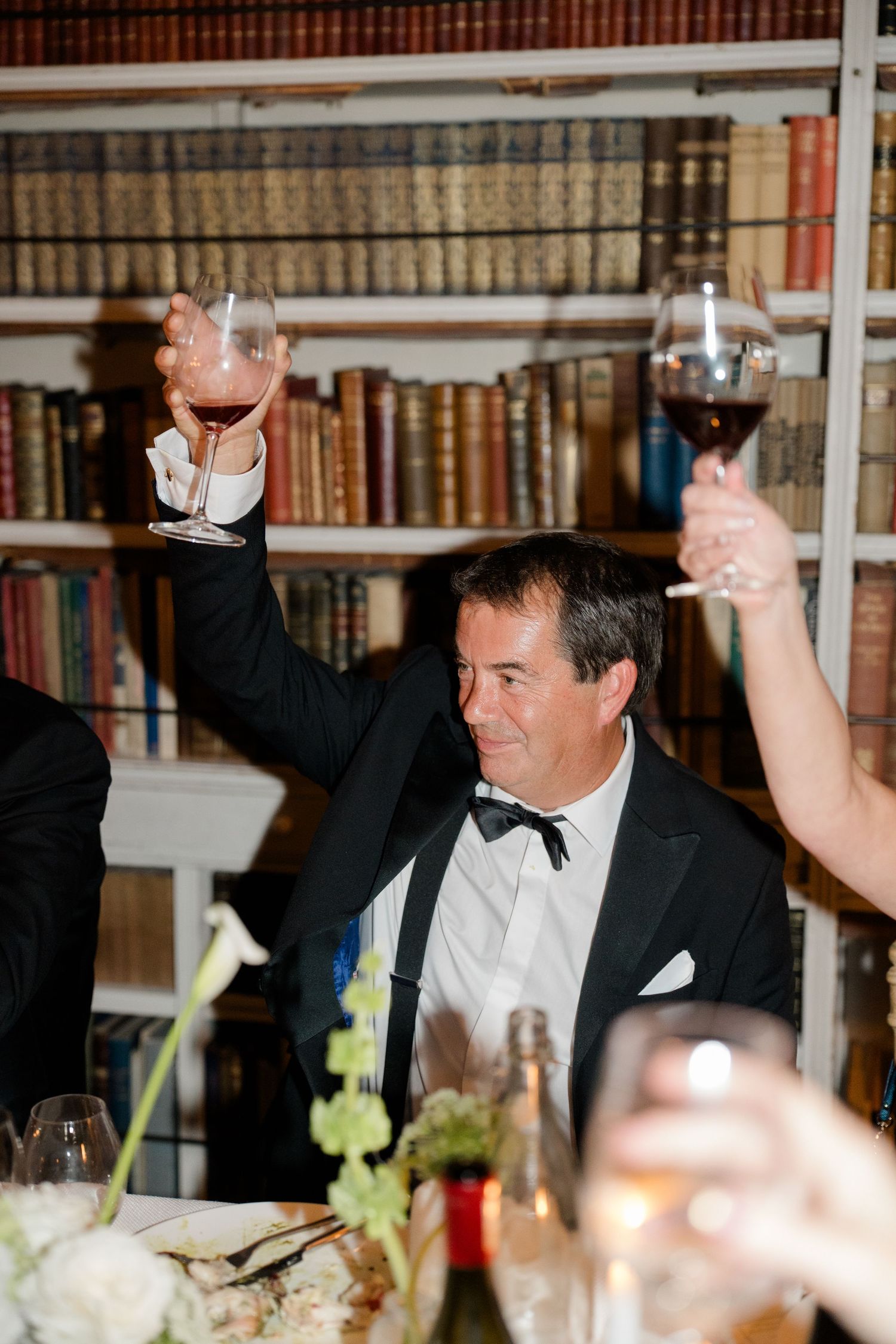 Guest in black tuxedo raises wine glass for a toast at wedding reception in a library setting.