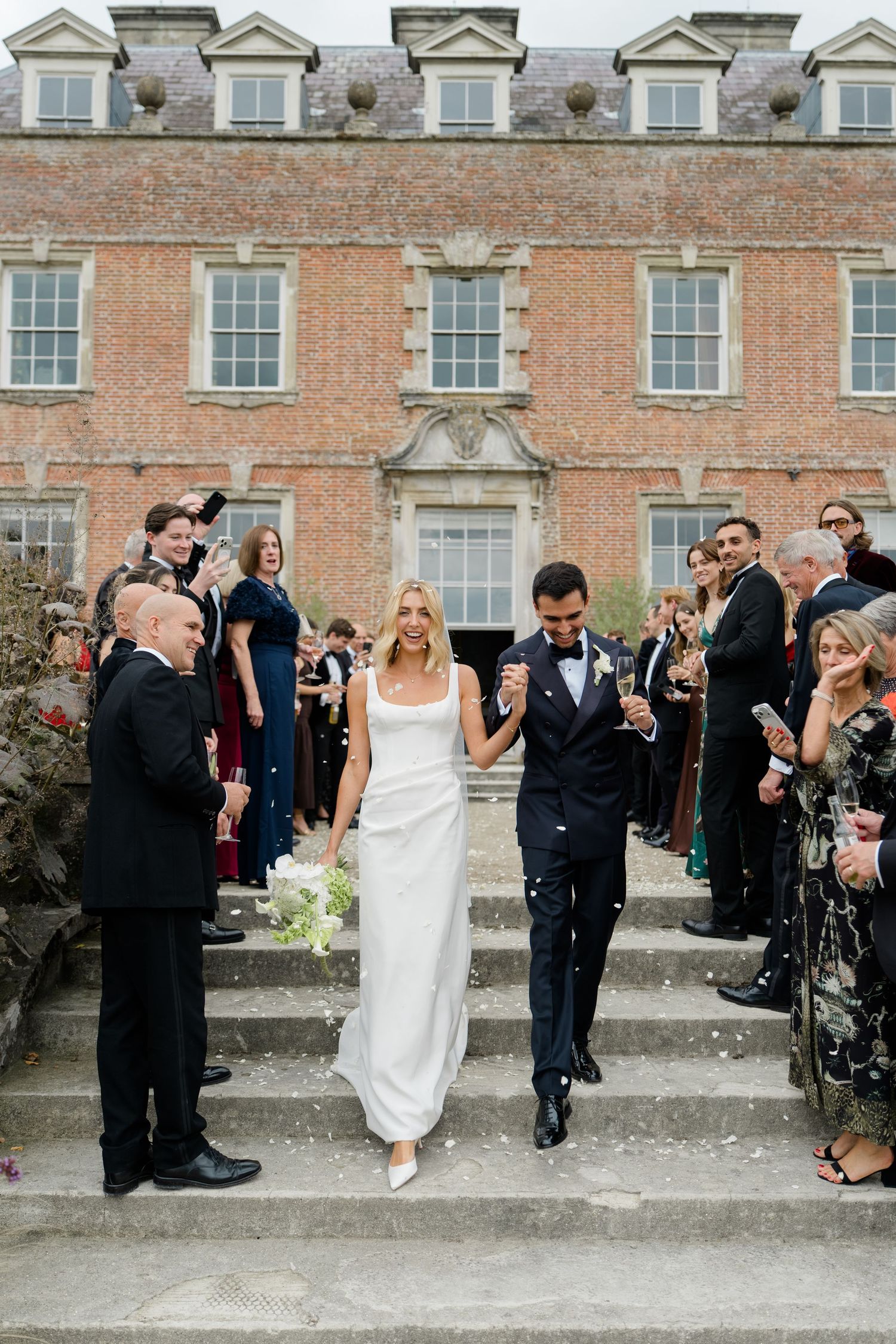Bride and groom walking down steps of grand English country house estate amid confetti shower.