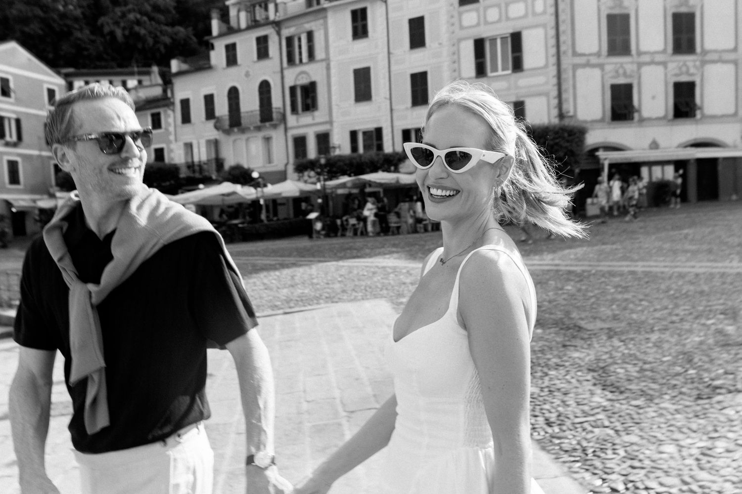 Black and white photo of a couple holding hands and walking in a European town square with historic buildings.