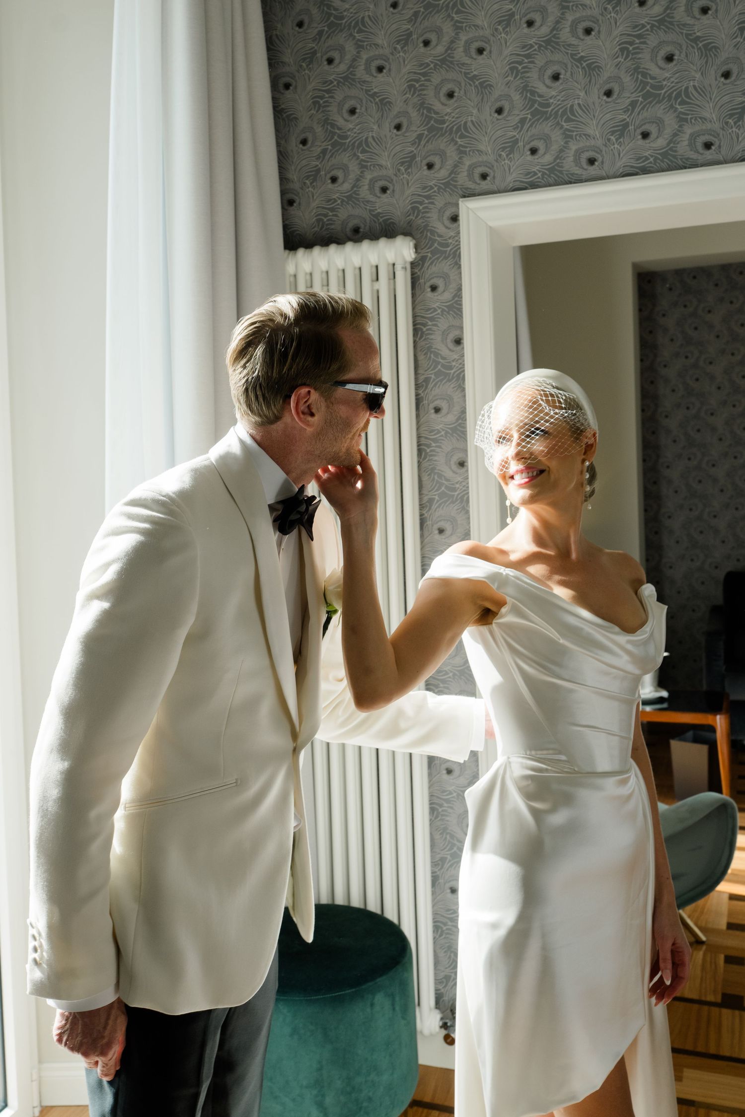 Groom in white tuxedo admires smiling bride in satin mini dress and birdcage veil in elegant hotel room.
