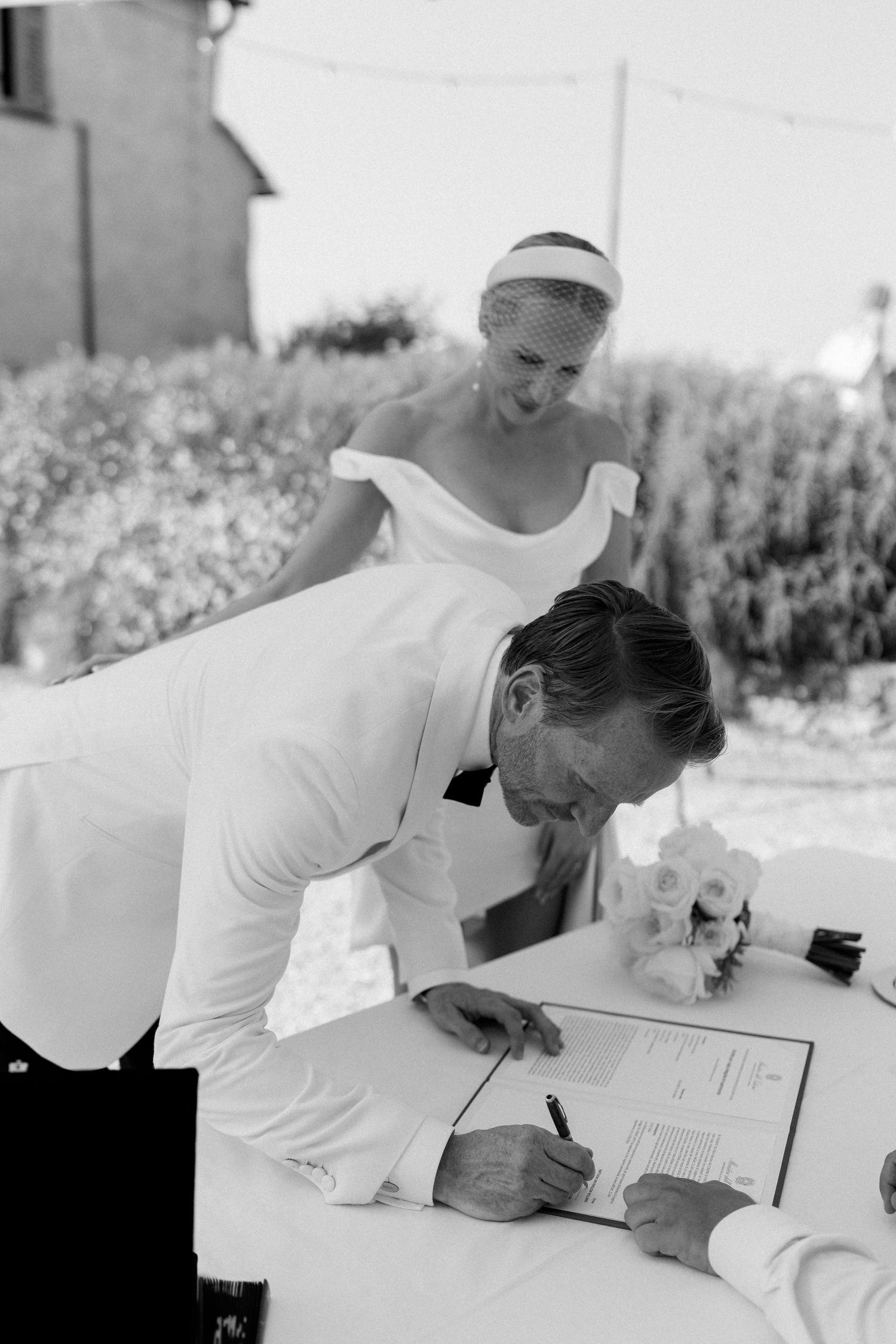 Groom in white jacket signs wedding certificate while smiling bride in off-shoulder dress and veil looks on.
