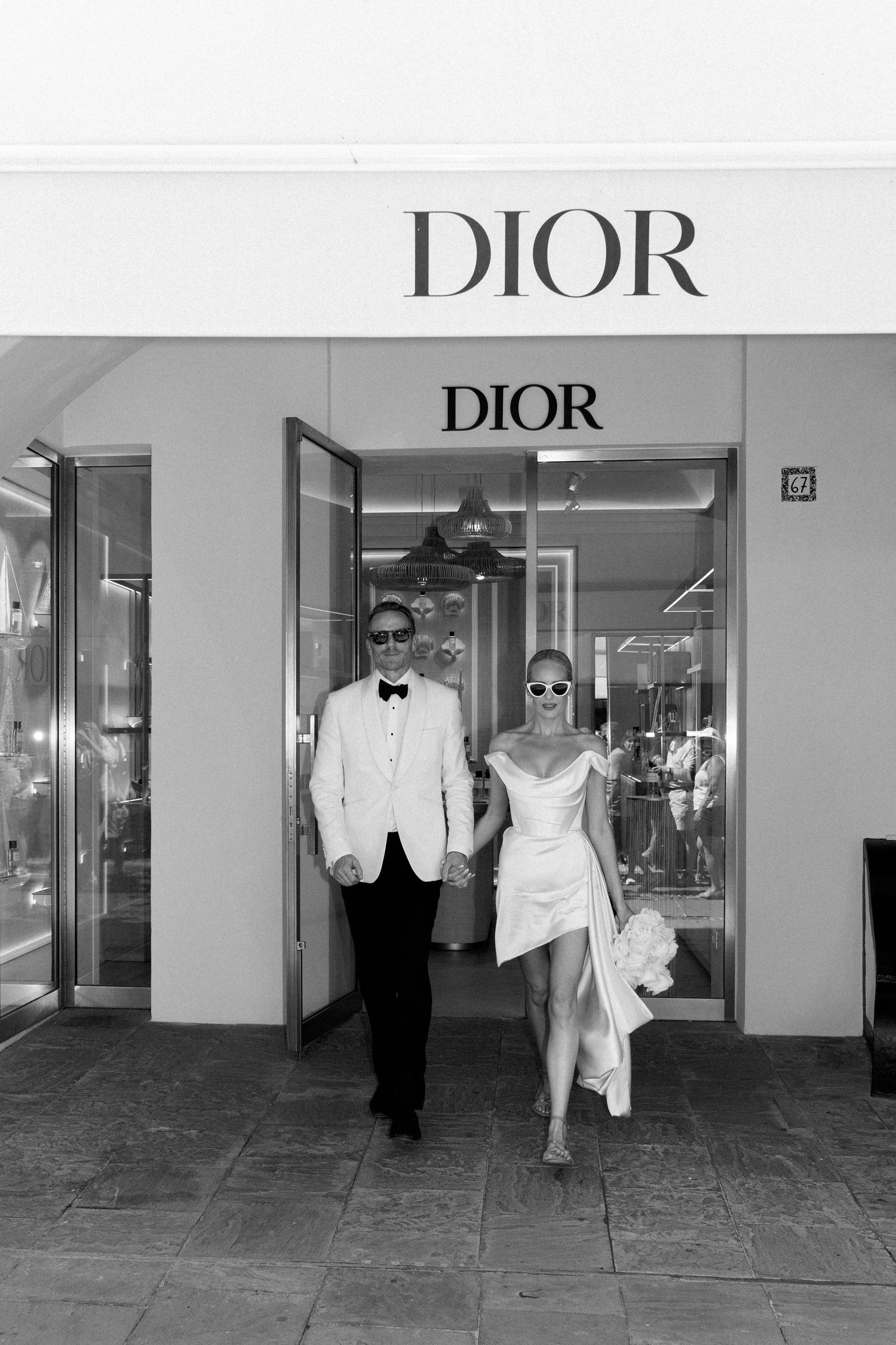 Elegant bride and groom in white wedding attire exiting a Dior boutique in black and white photo.