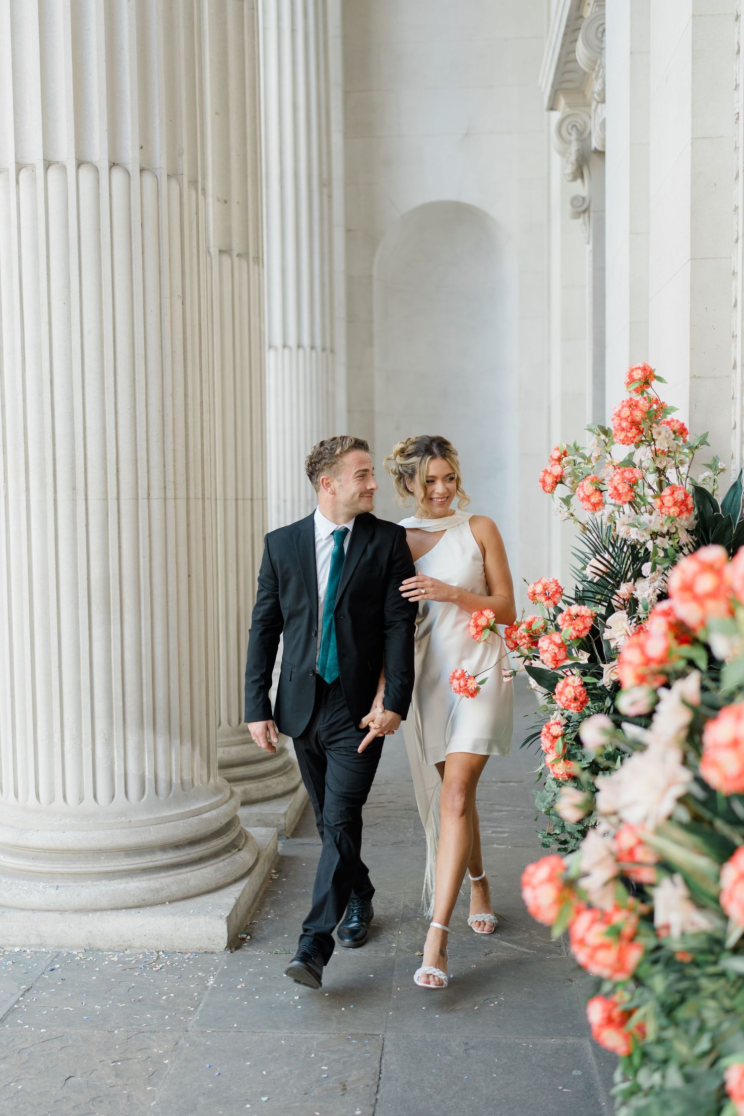 Couple walks playfully between white columns with coral flowers in foreground.