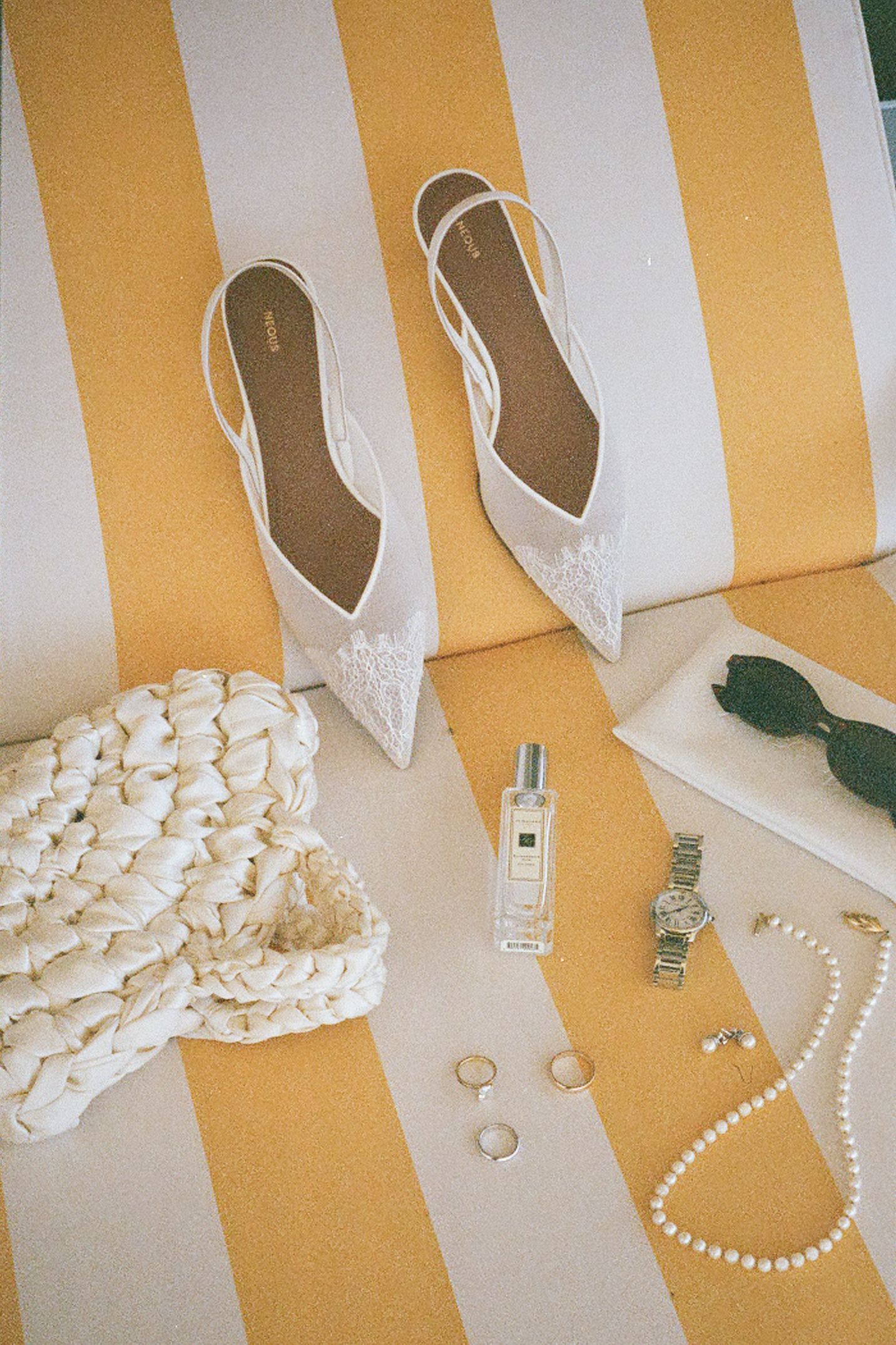 Wedding accessories including pearl necklaces, white shoes, and jewelry arranged in a row on a beige and white striped surface.