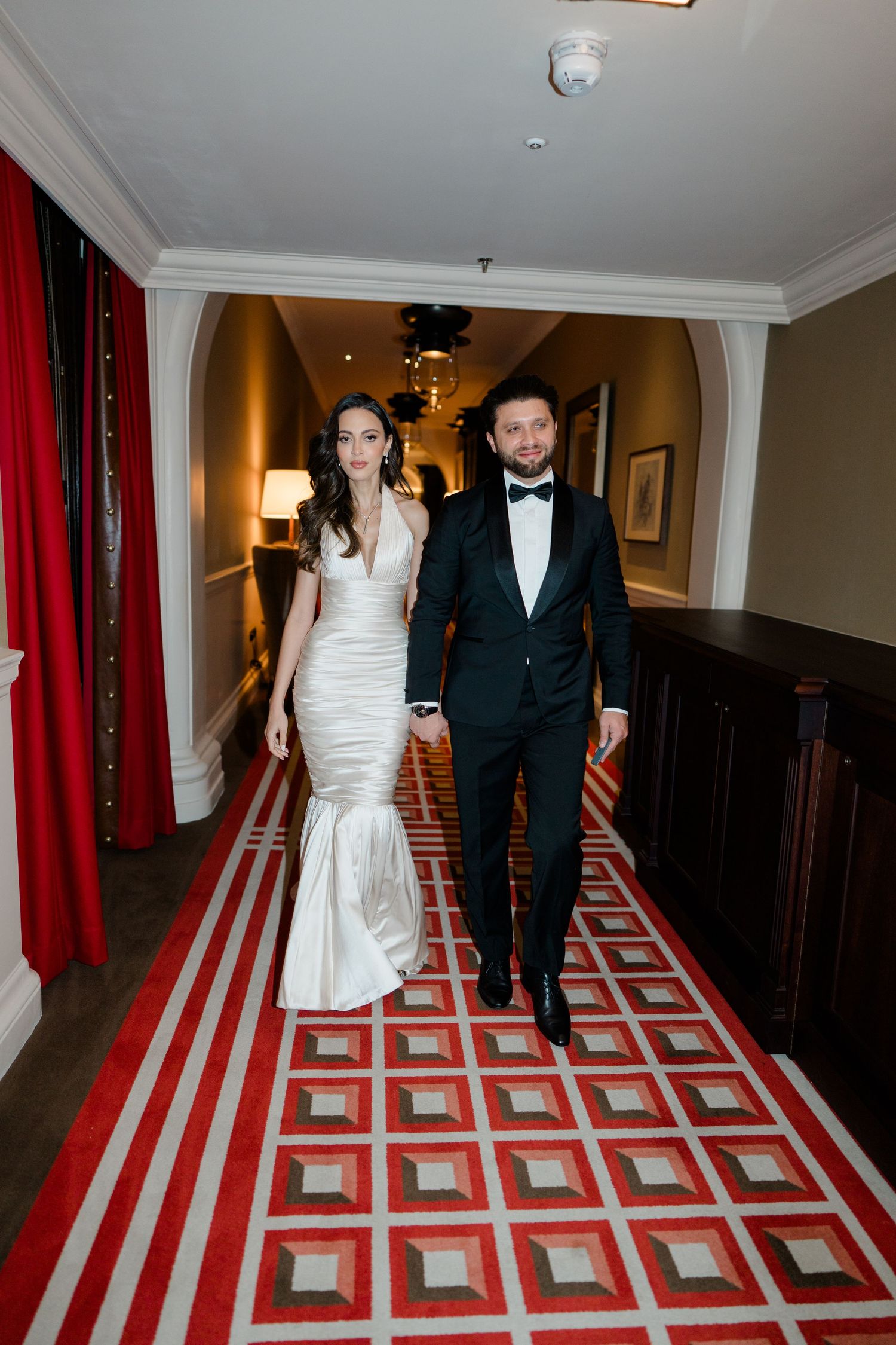 Couple in formal attire walking down a hotel hallway with distinctive red and white geometric carpet pattern.