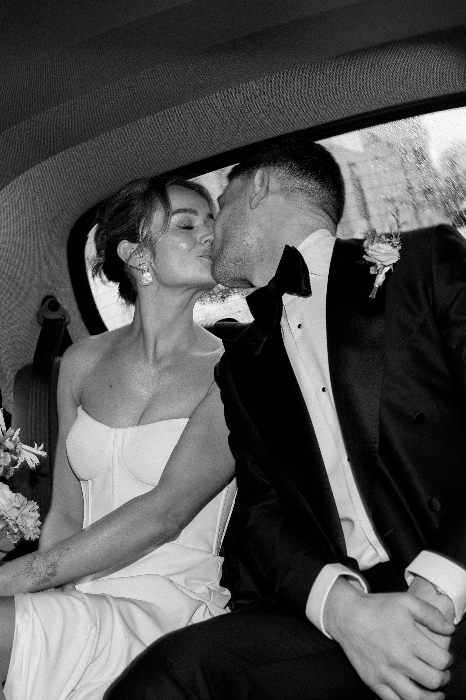 A romantic black and white photo of newlyweds sharing a kiss in the back of a car after their wedding ceremony.