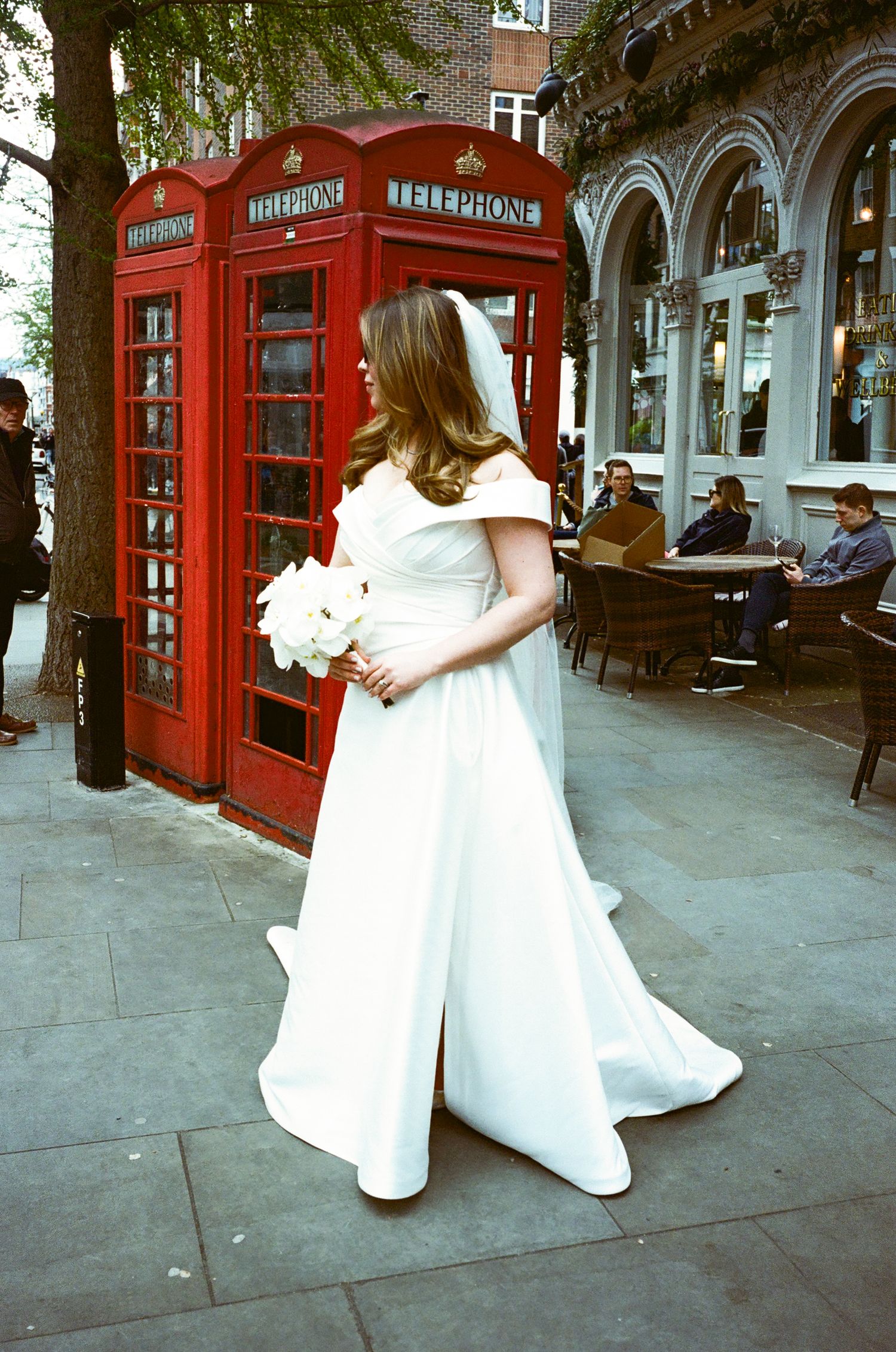 Someone in a white wedding dress holds a bouquet while standing next to an iconic red London telephone booth on a city street.