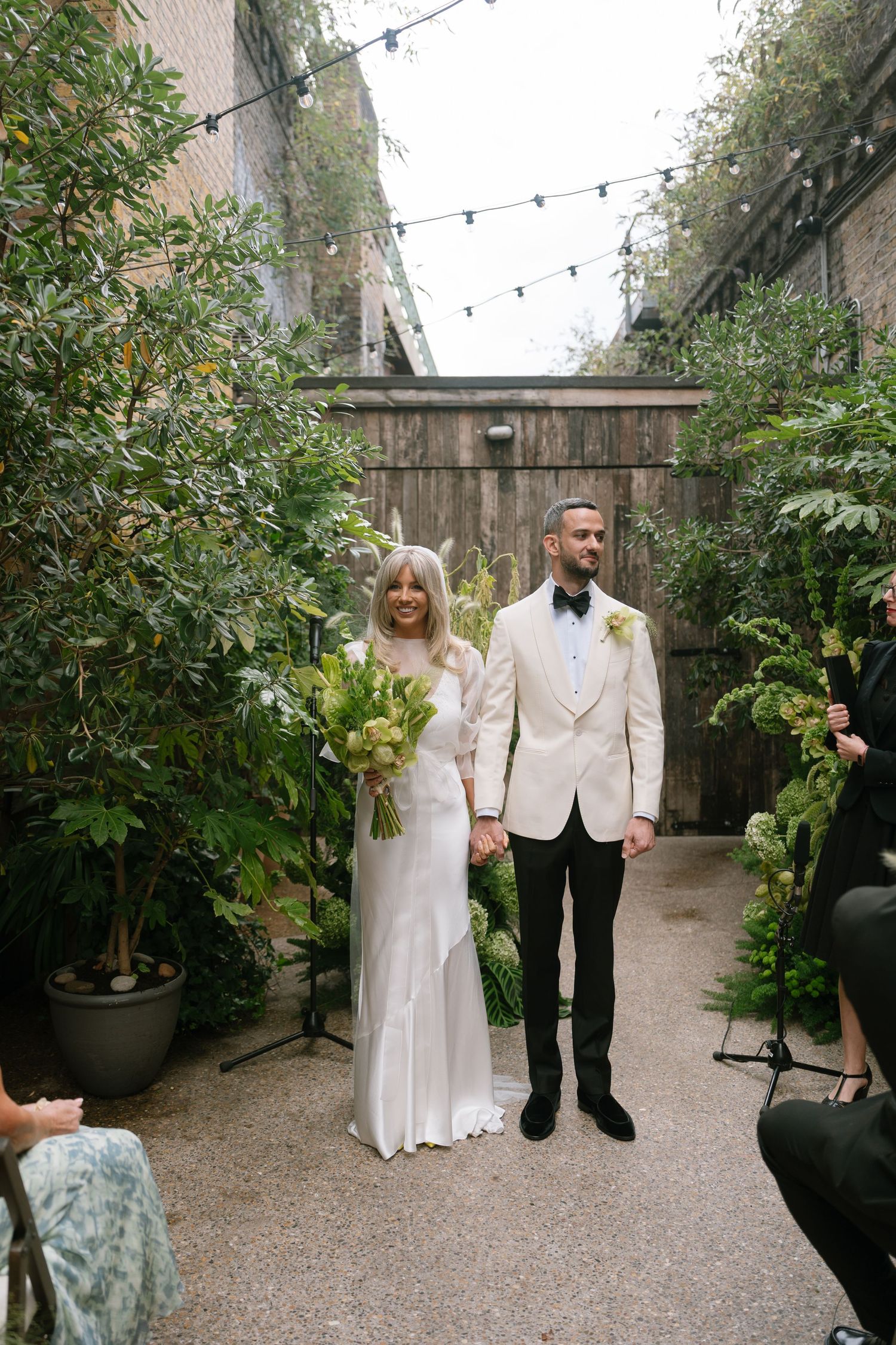 Wedding couple poses in a garden courtyard with string lights and greenery.