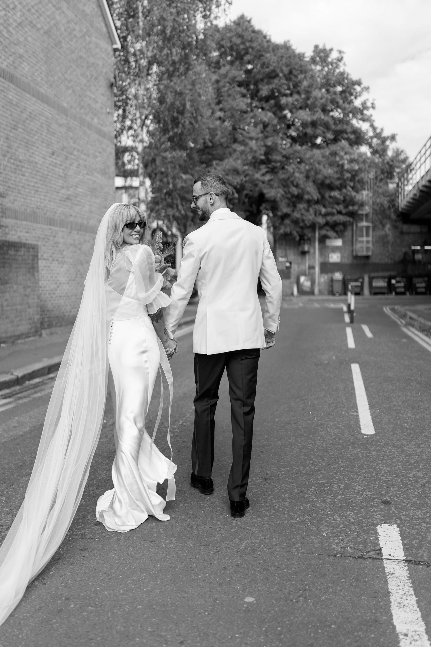 A couple in wedding attire walks along a paved path in black and white.