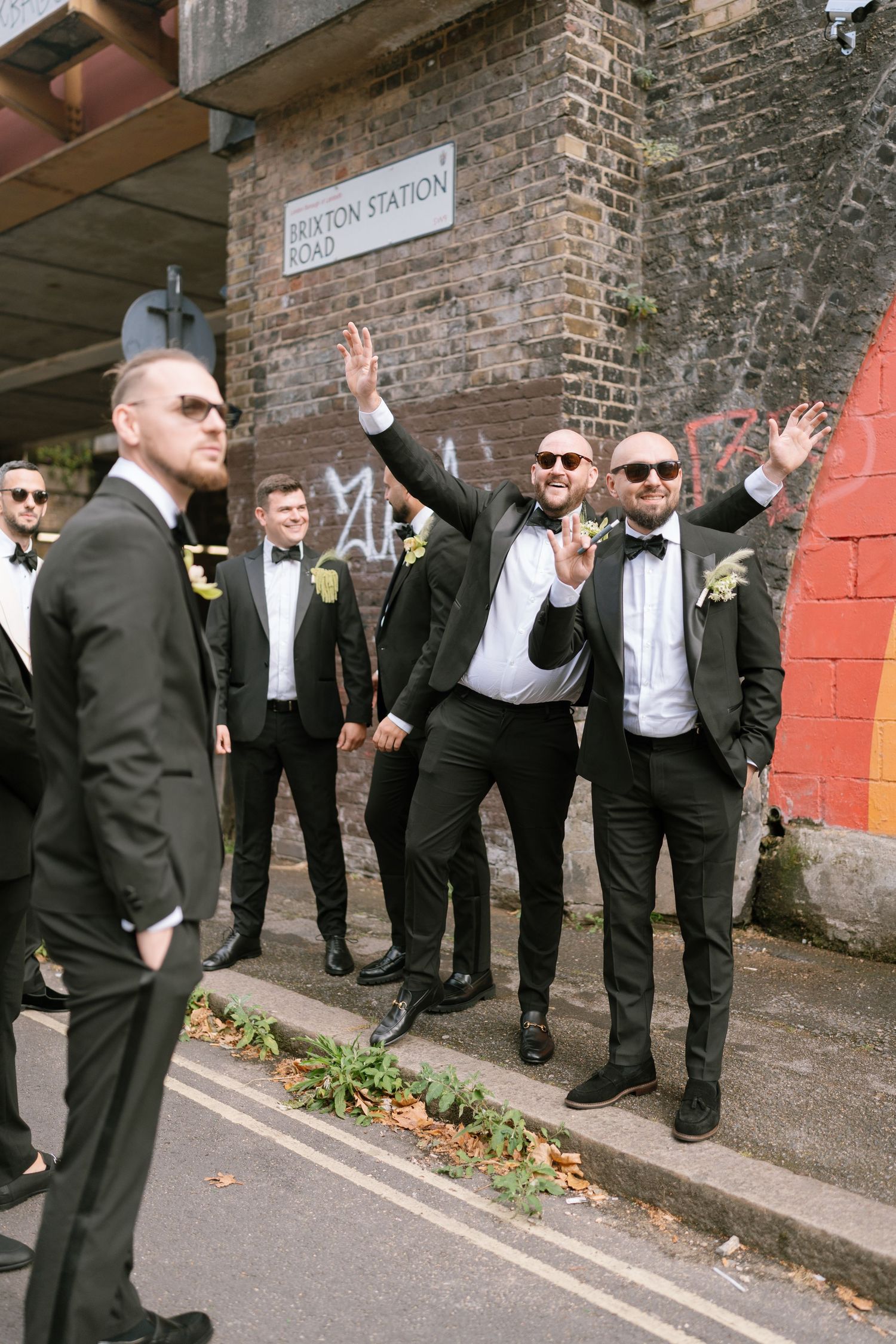 Groomsmen in black suits pose playfully outside brick building with street sign.