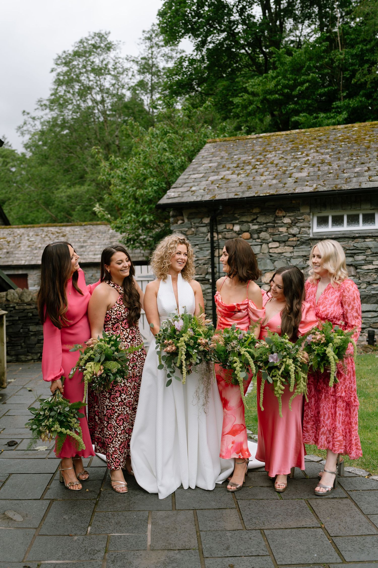 Wedding party wearing pink and white dresses poses together against stone wall with greenery bouquets.
