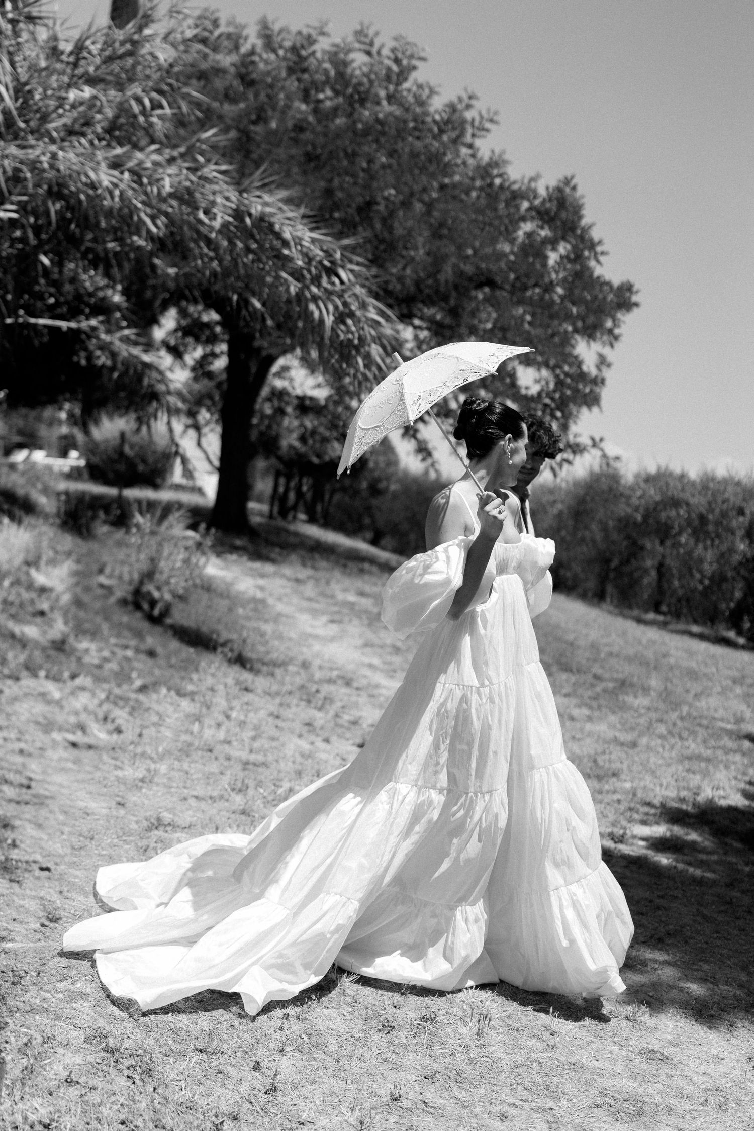A black and white photo of a figure in a flowing white dress holding a parasol in a garden setting.