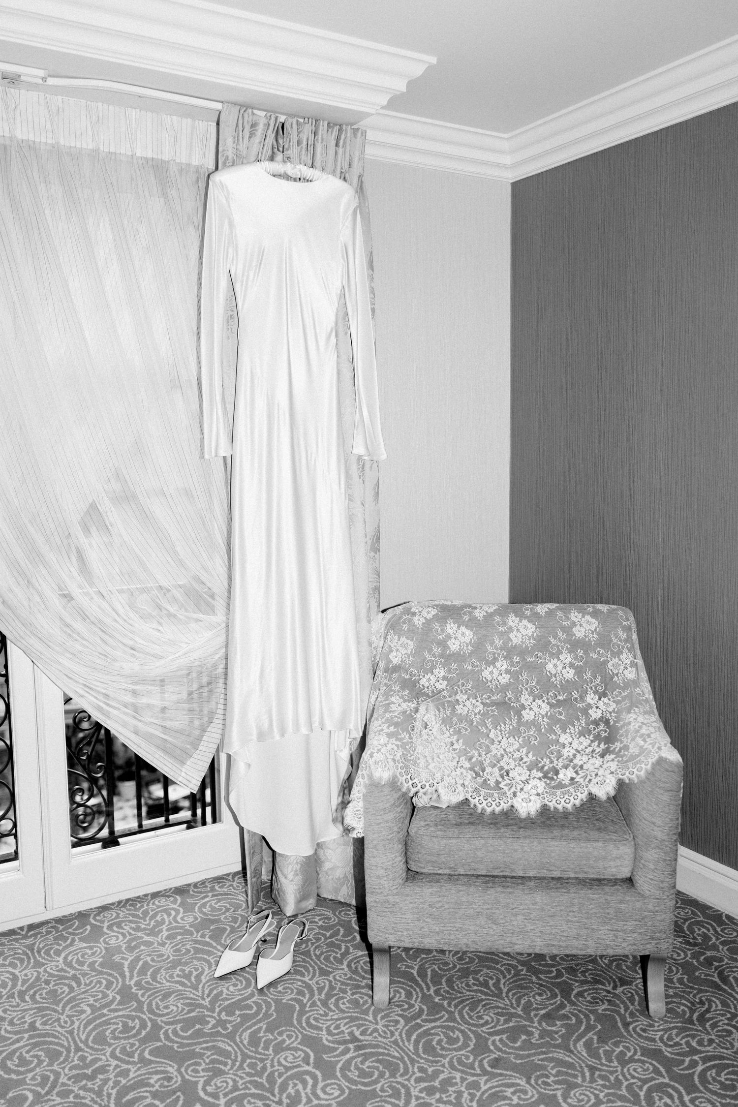 Elegant white wedding dress hanging in a room with sheer curtains and a vintage armchair with lace doily.