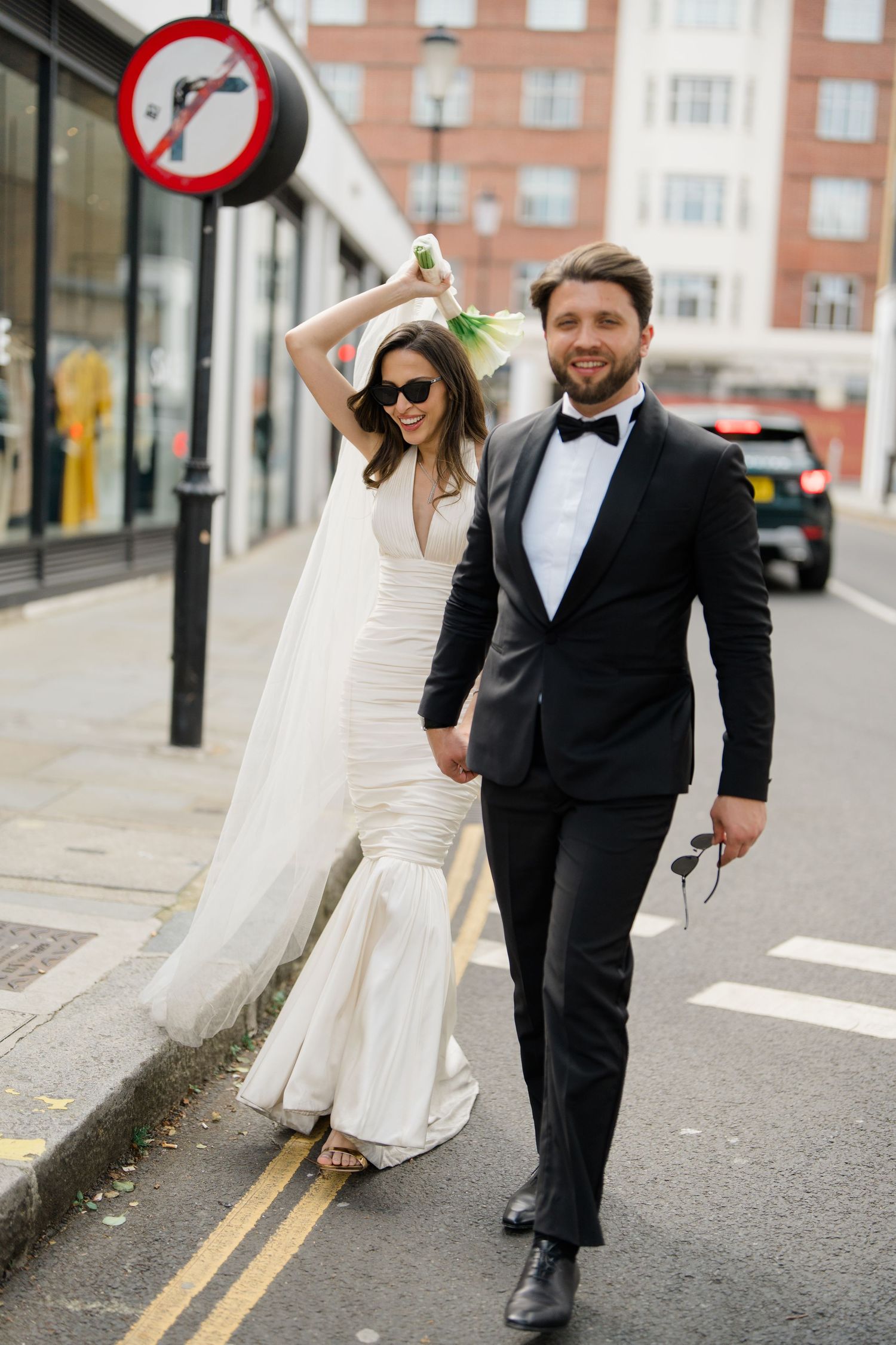 A bride in a white gown and groom in a tuxedo walk hand-in-hand on a London street, bride raising bouquet joyfully.