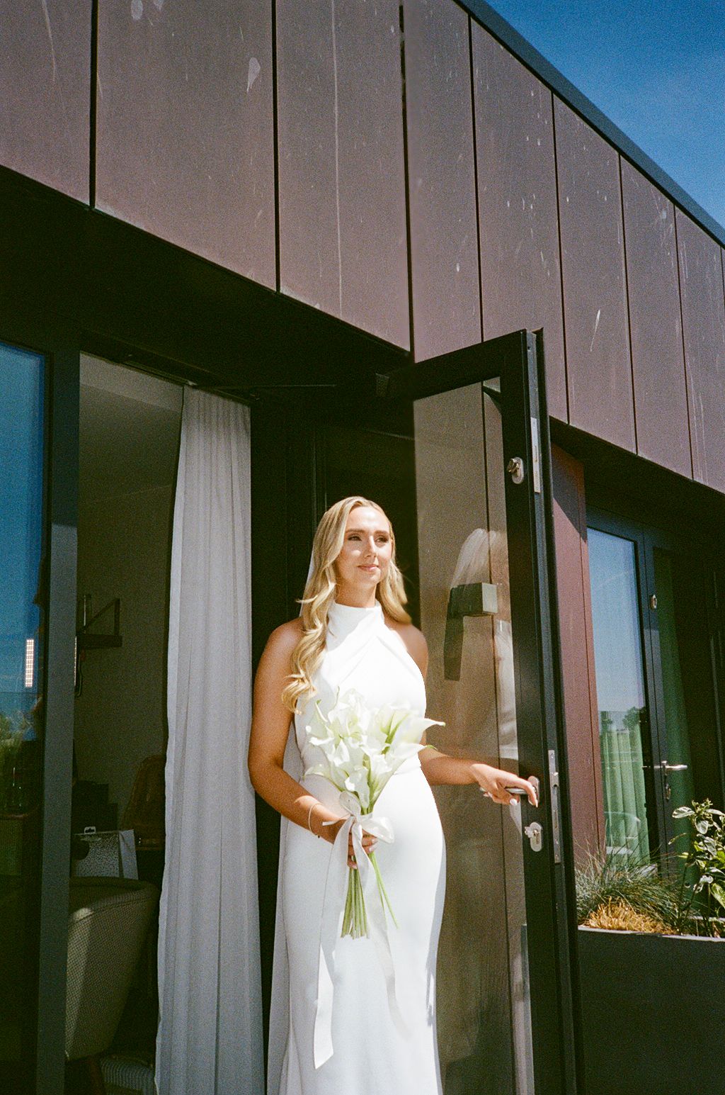 Bride in white halter gown holding calla lily bouquet standing at modern dark wood building entrance on sunny day.