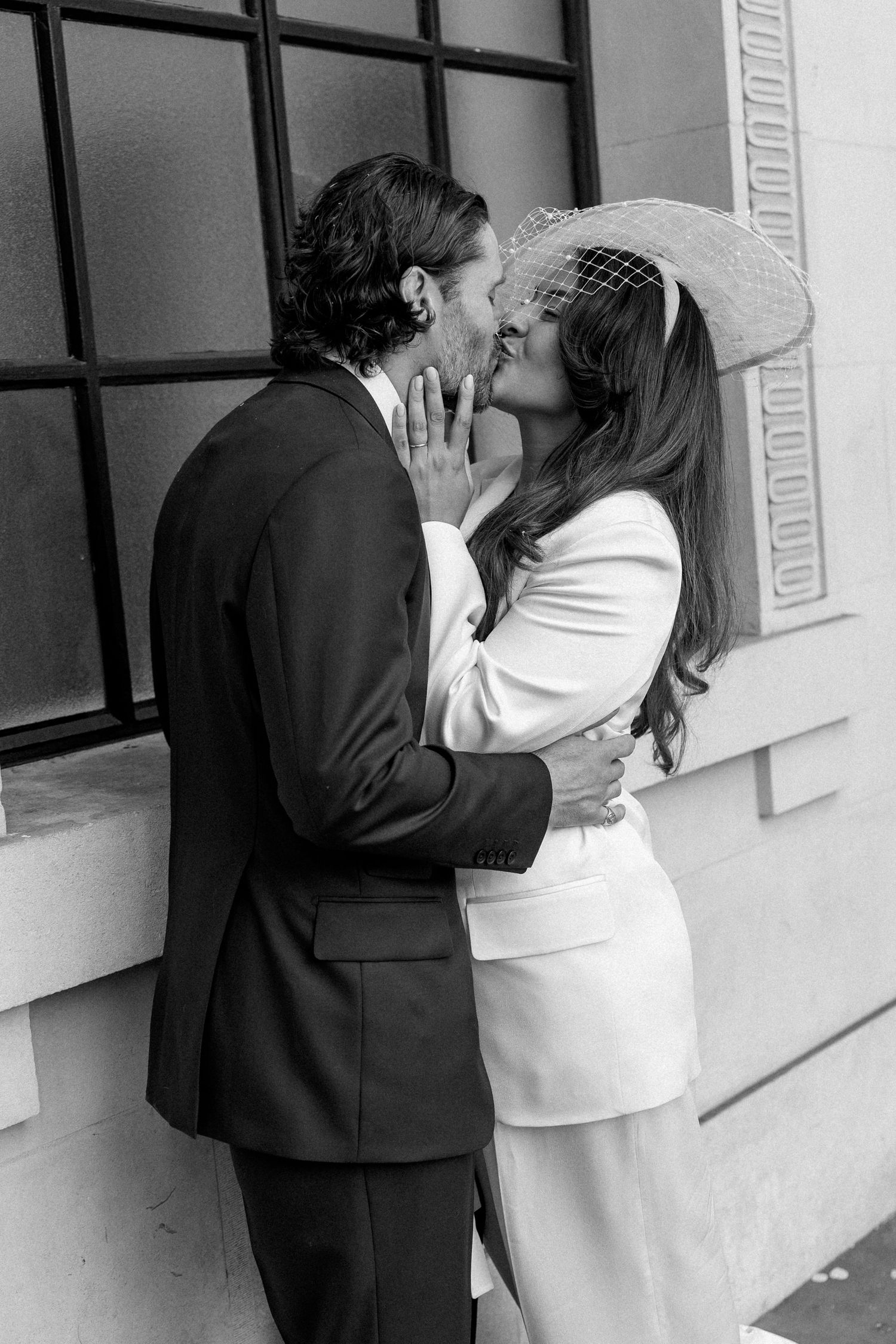 Newlyweds sharing a romantic kiss, bride in white blazer and birdcage hat embracing suited groom.