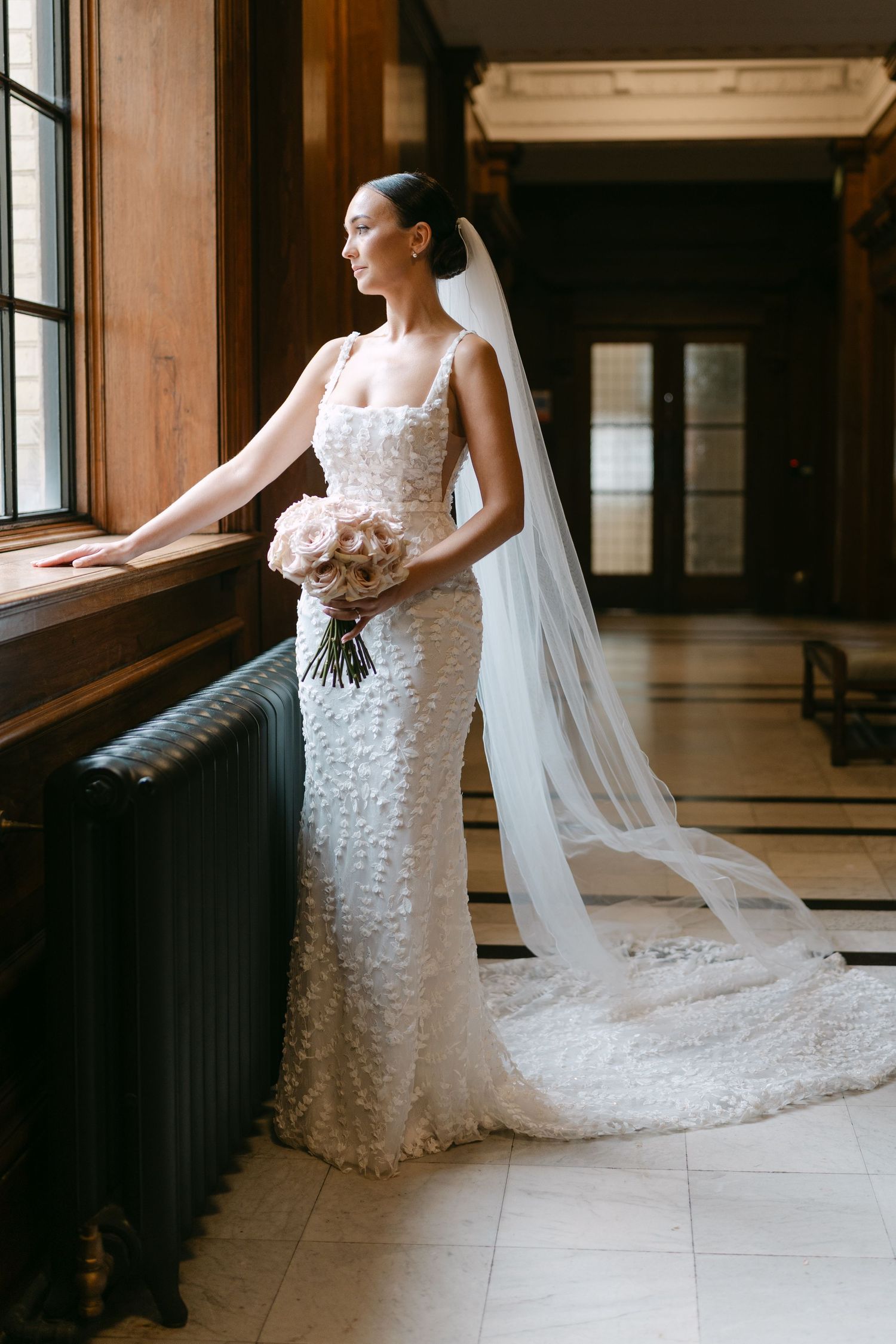Bride in lace gown with flowing veil stands by window in elegant venue.