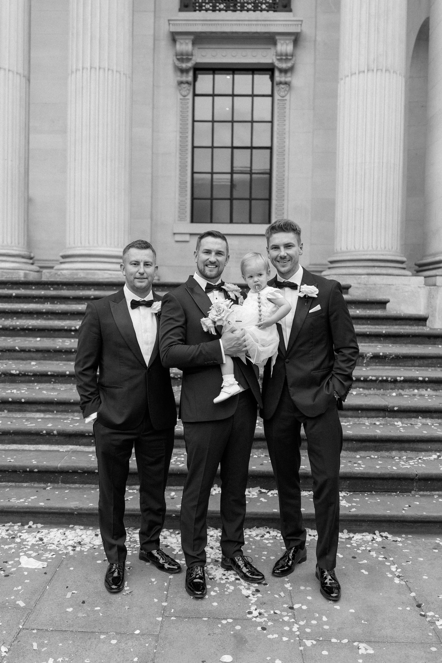 Groomsmen in black tuxedos pose casually on stone steps with flower petals.