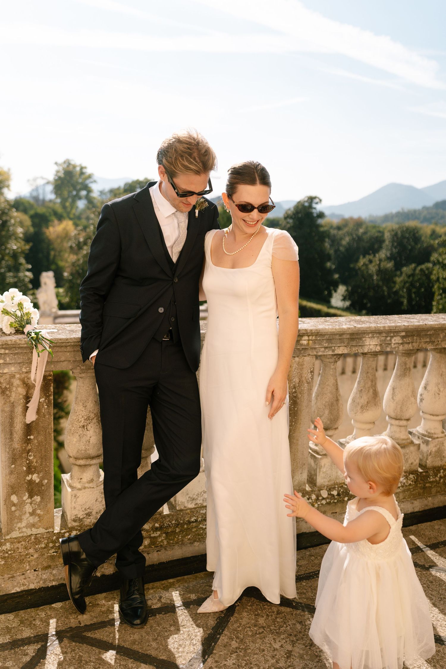Wedding couple in sunglasses share a moment with small child on stone balcony.