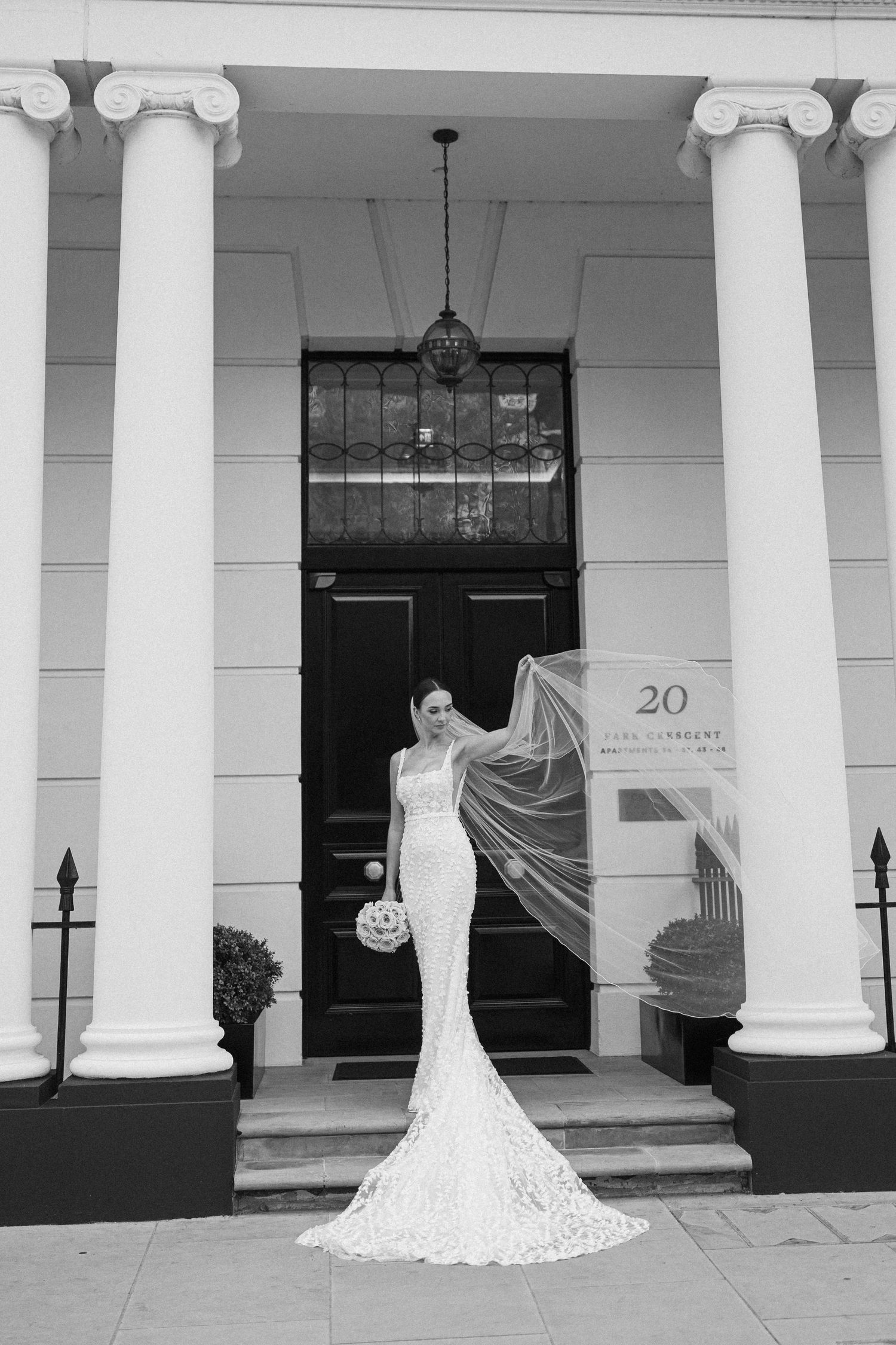 A bride in a fitted lace gown poses with flowing veil in front of a black door with white columns.