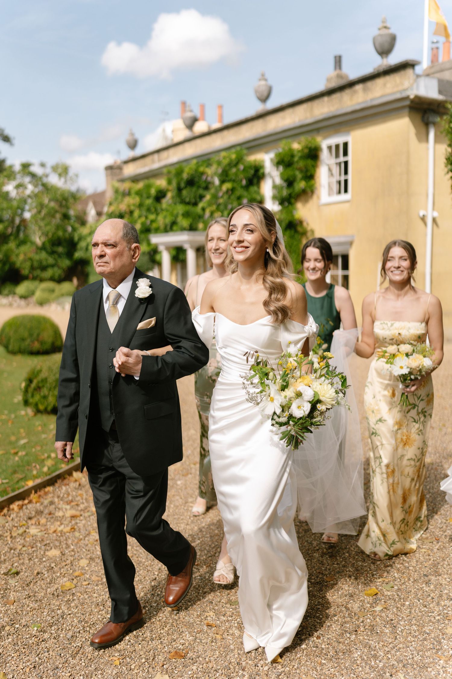 Wedding party walking along gravel path outside elegant yellow country manor house.