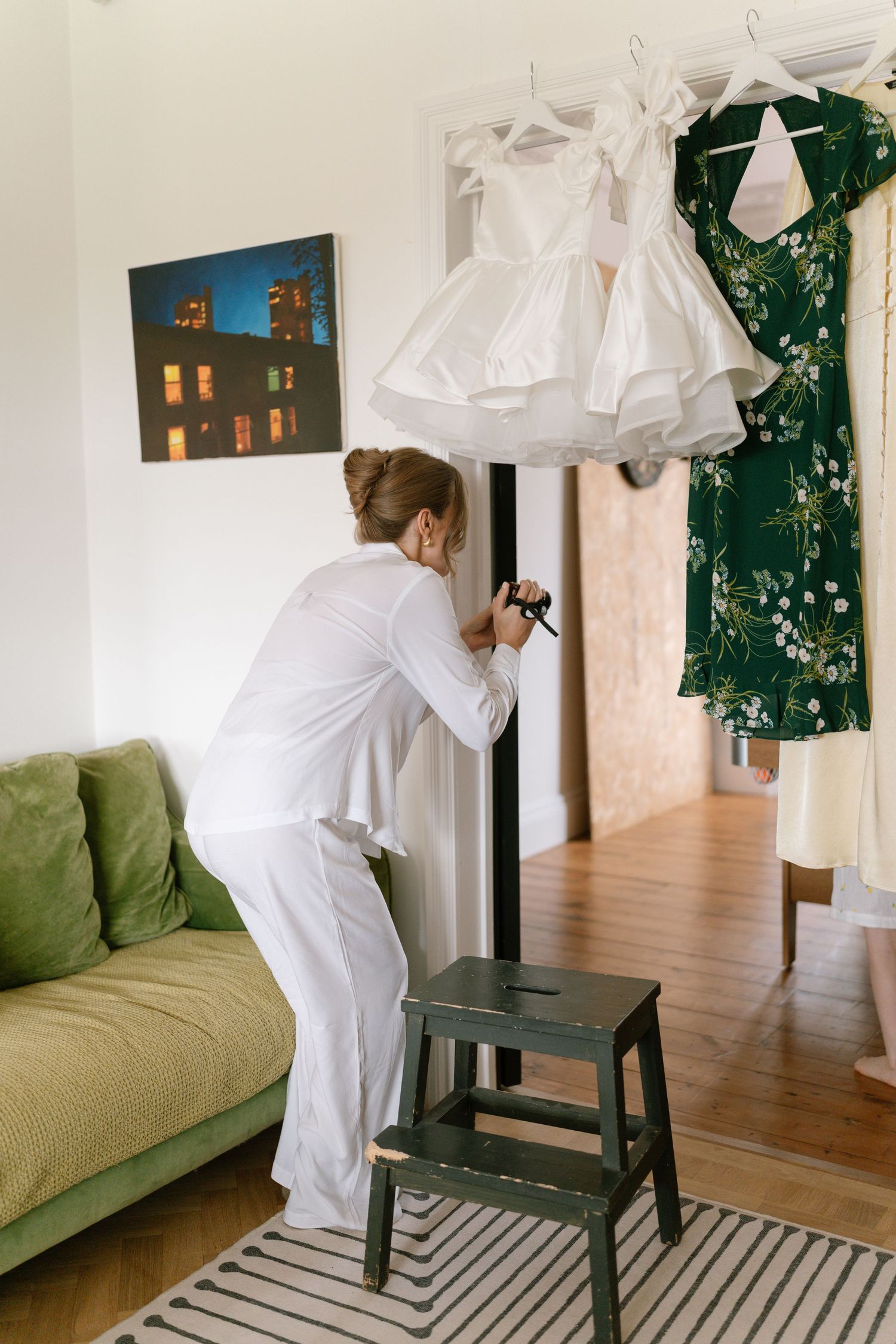 Person in white photographing hanging dresses against wall with green couch.