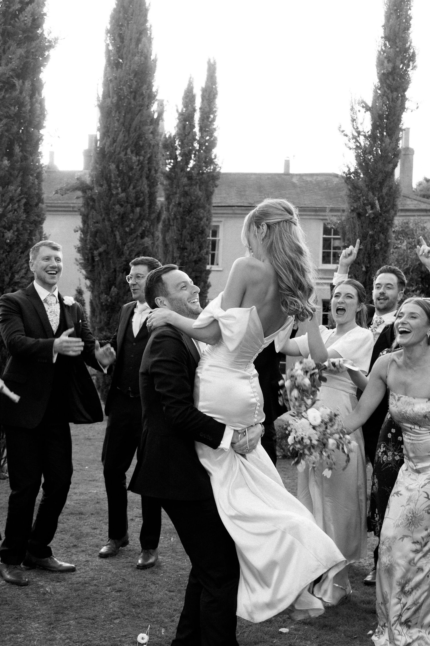 Wedding party members share joyful candid moments outdoors with tall cypress trees in the background.