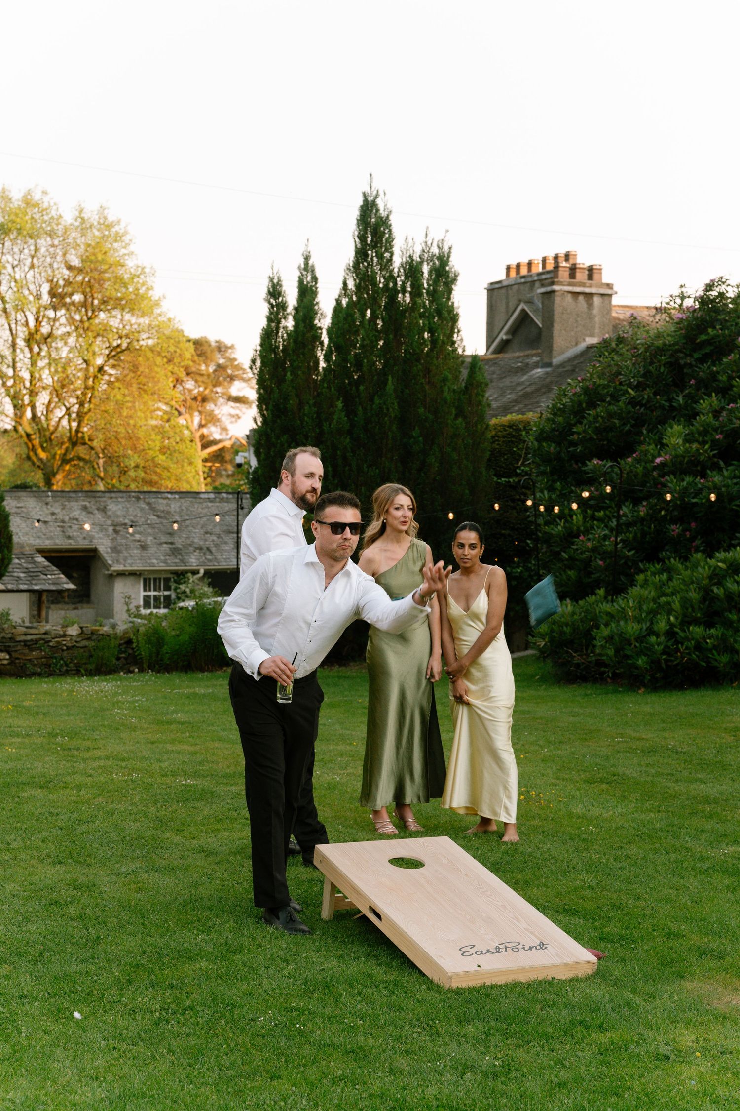 Wedding party members enjoy a game of cornhole on a lawn during golden hour.