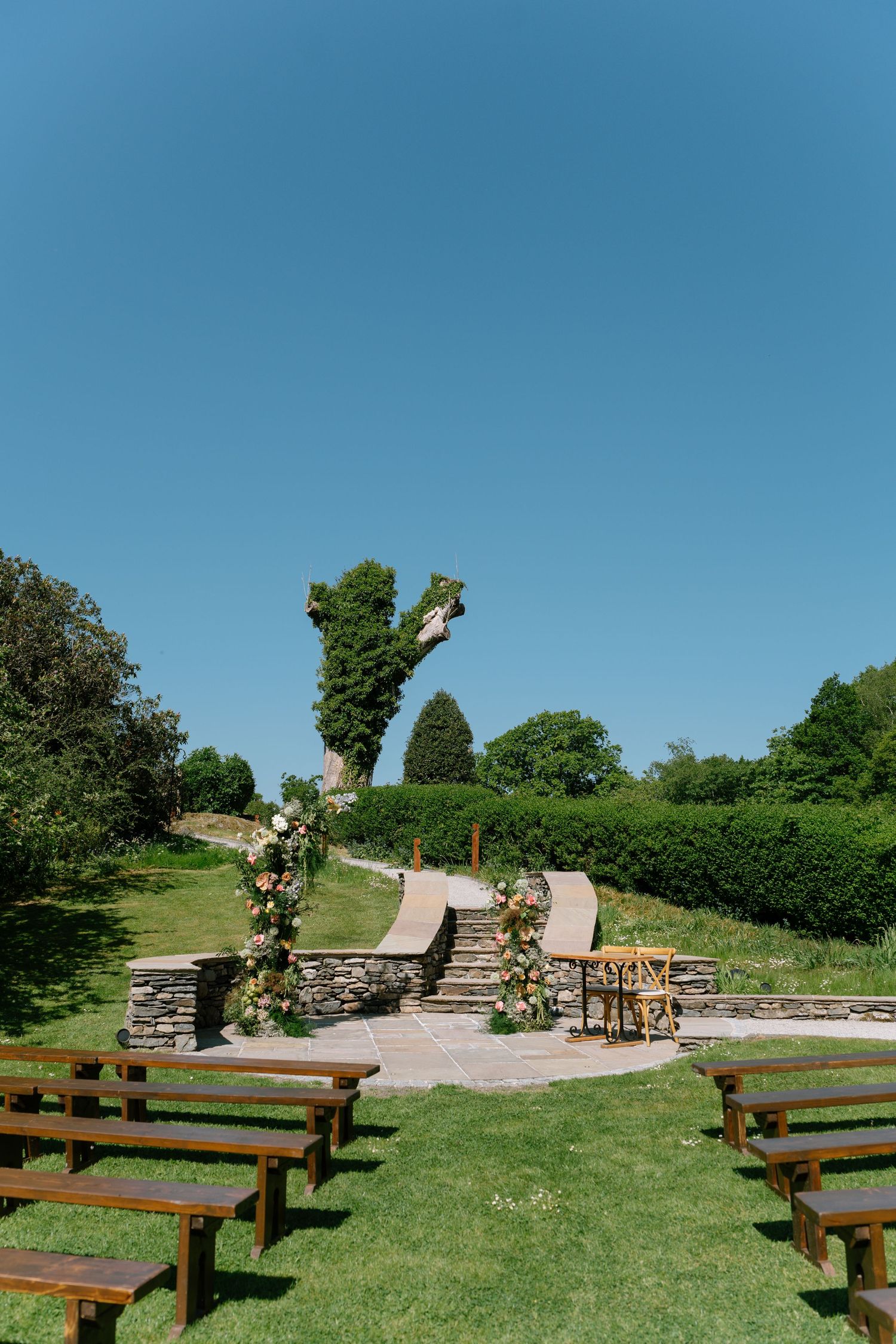 Rustic outdoor ceremony space with wooden benches arranged in rows facing a stone altar area.