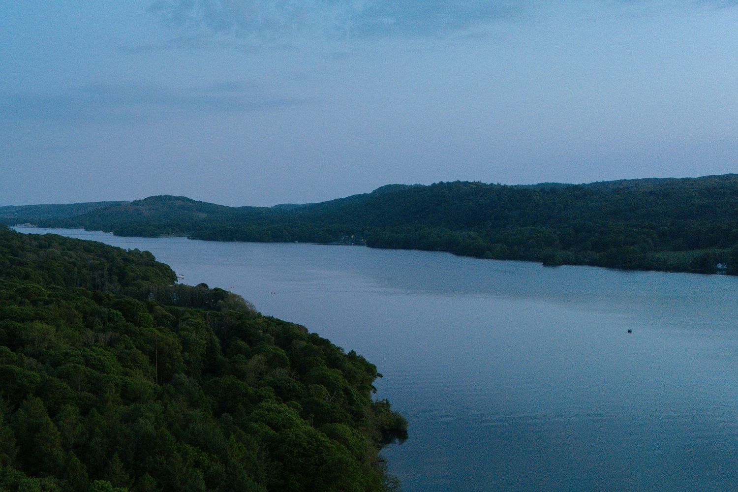 Panoramic view of a serene lake surrounded by forested hills under blue skies.