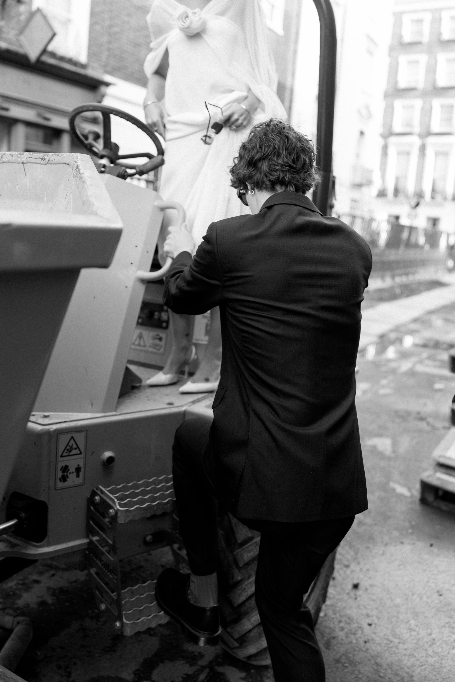 Black and white photo of couple posing near industrial equipment in urban setting.