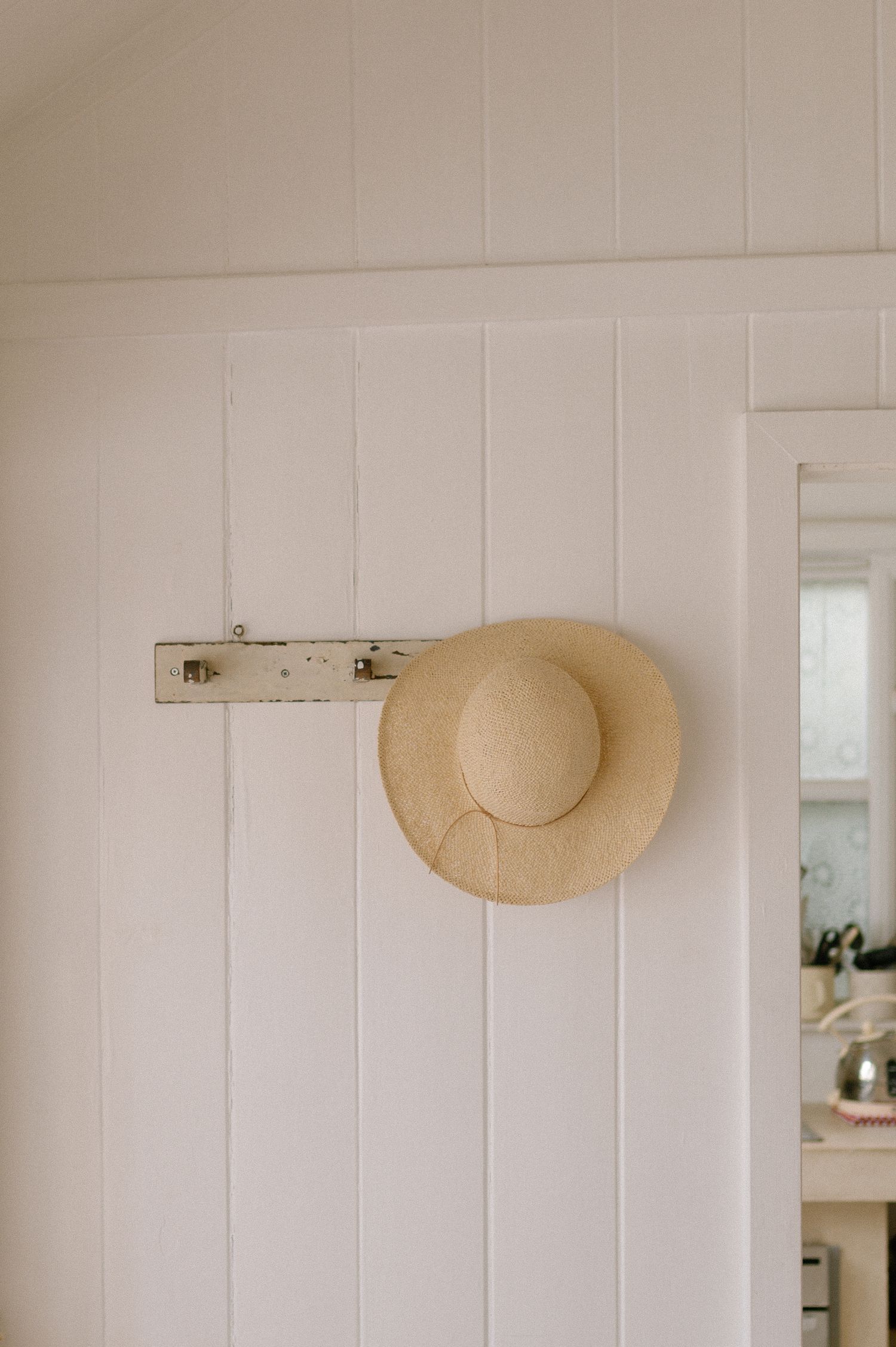 A straw sun hat hangs on a white wooden wall in a bright, airy room.