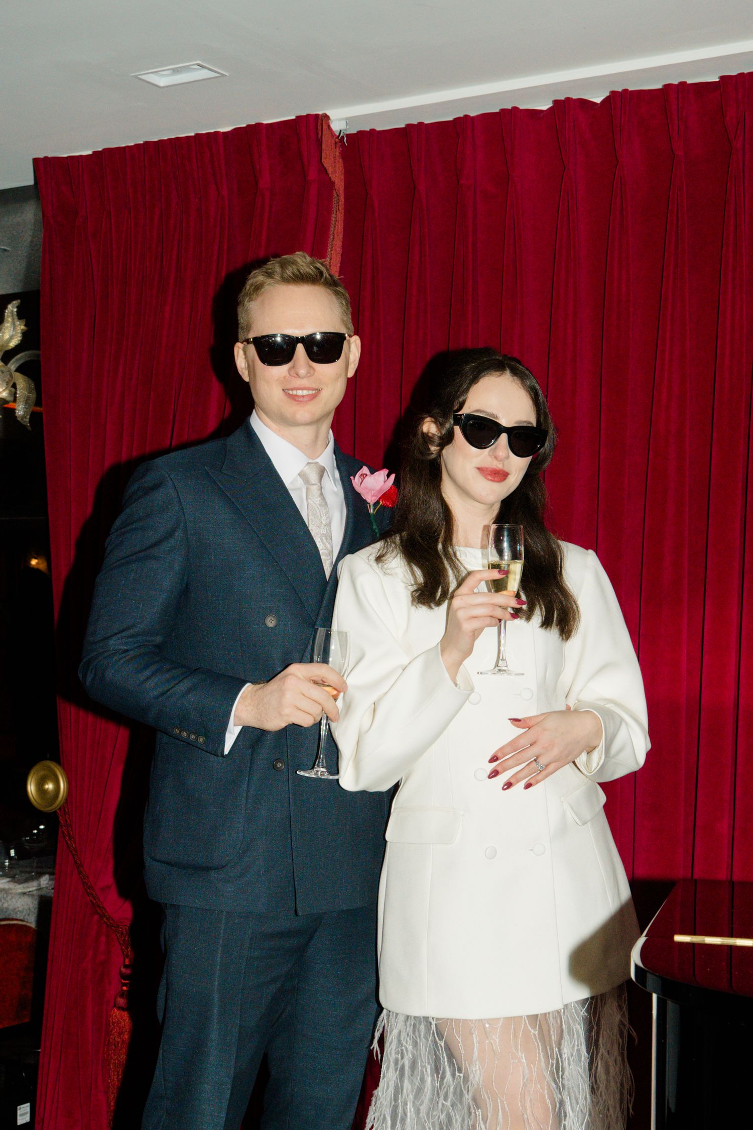 Bride and groom in sunglasses toasting champagne before red velvet curtain backdrop.