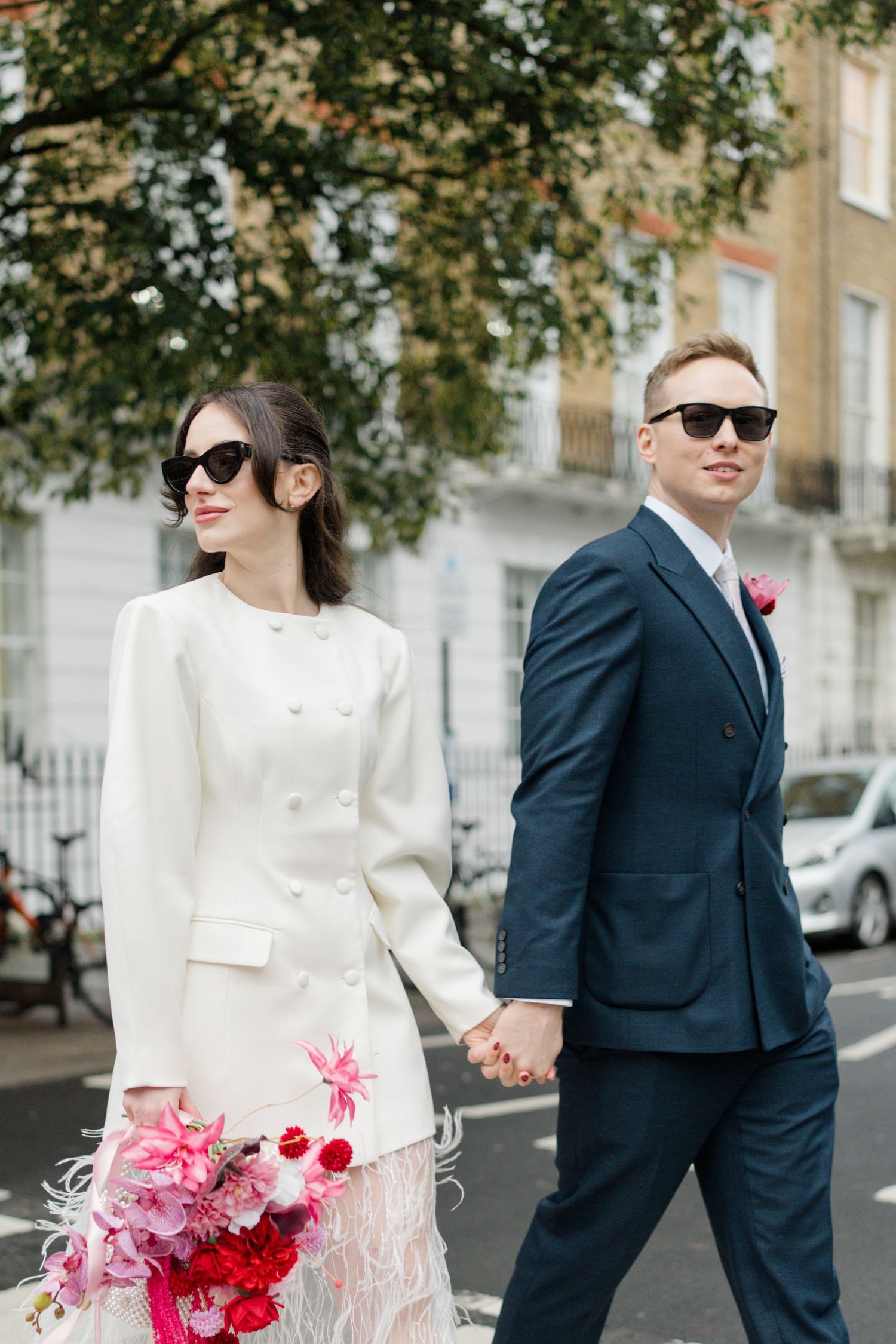 Stylish bride and groom in sunglasses hold hands crossing street, bride carries vibrant pink bouquet.
