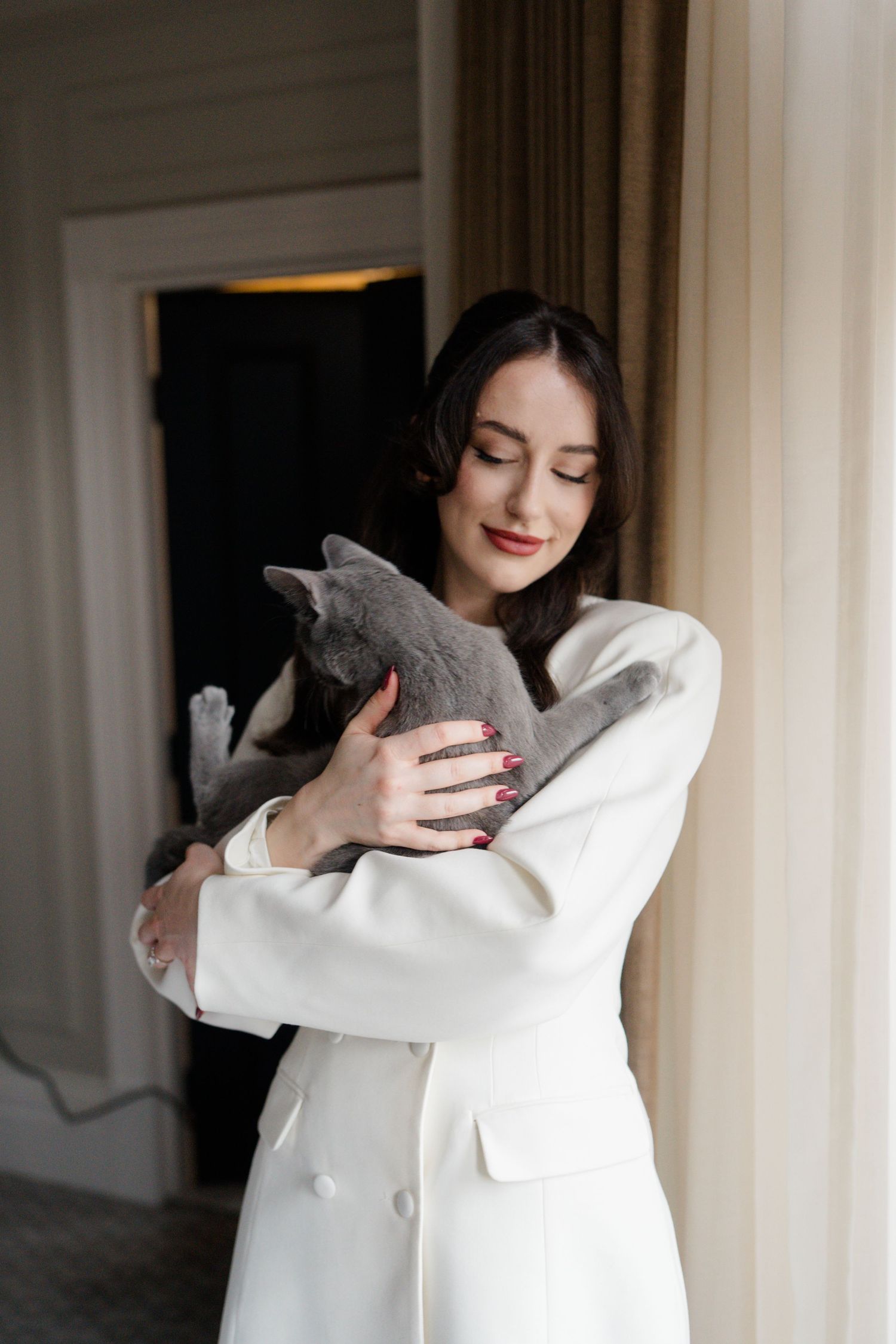Young woman in white blazer tenderly hugging a grey Russian Blue cat near a window.