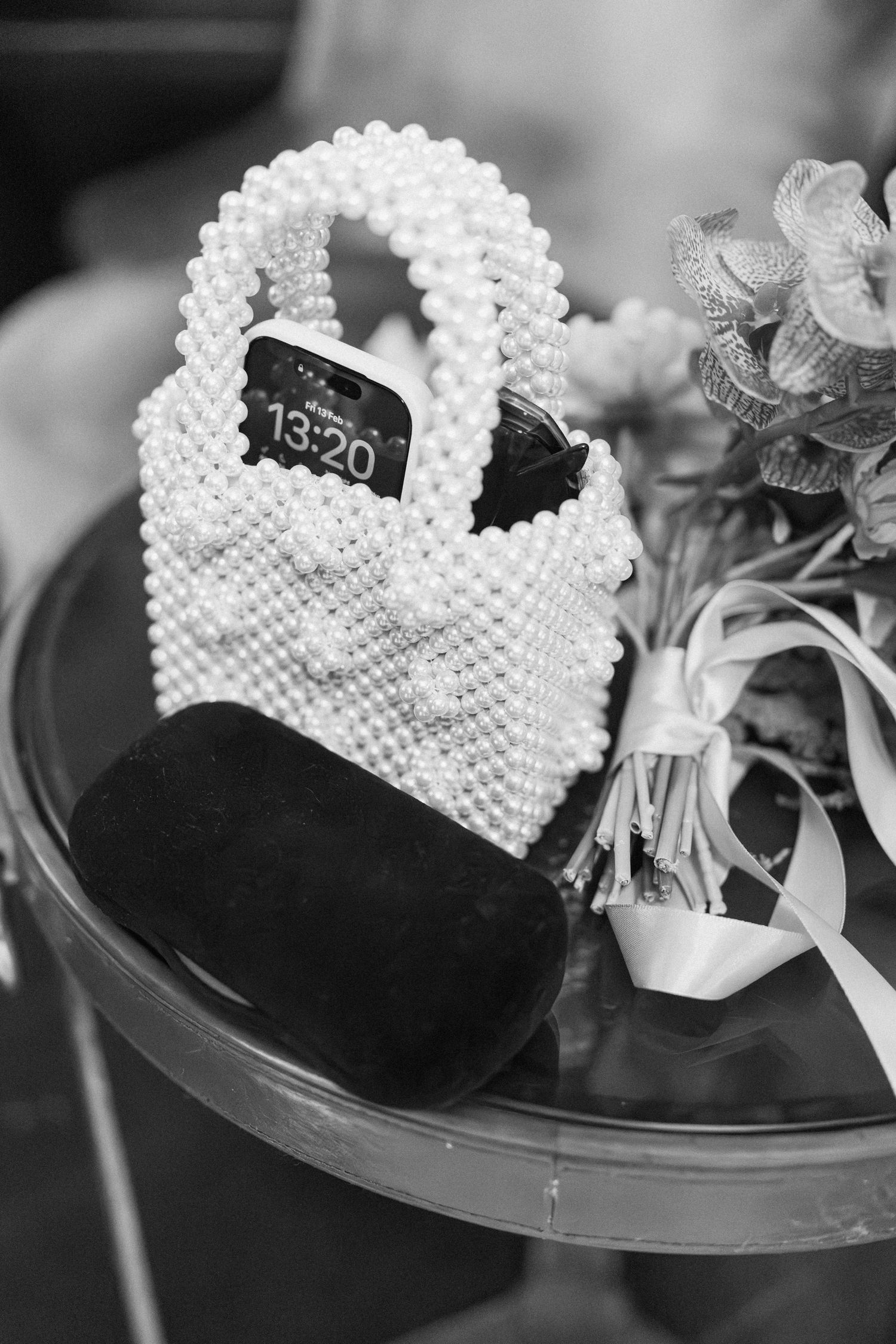Pearl beaded bridal handbag with phone showing 13:20 beside a bouquet in black and white.