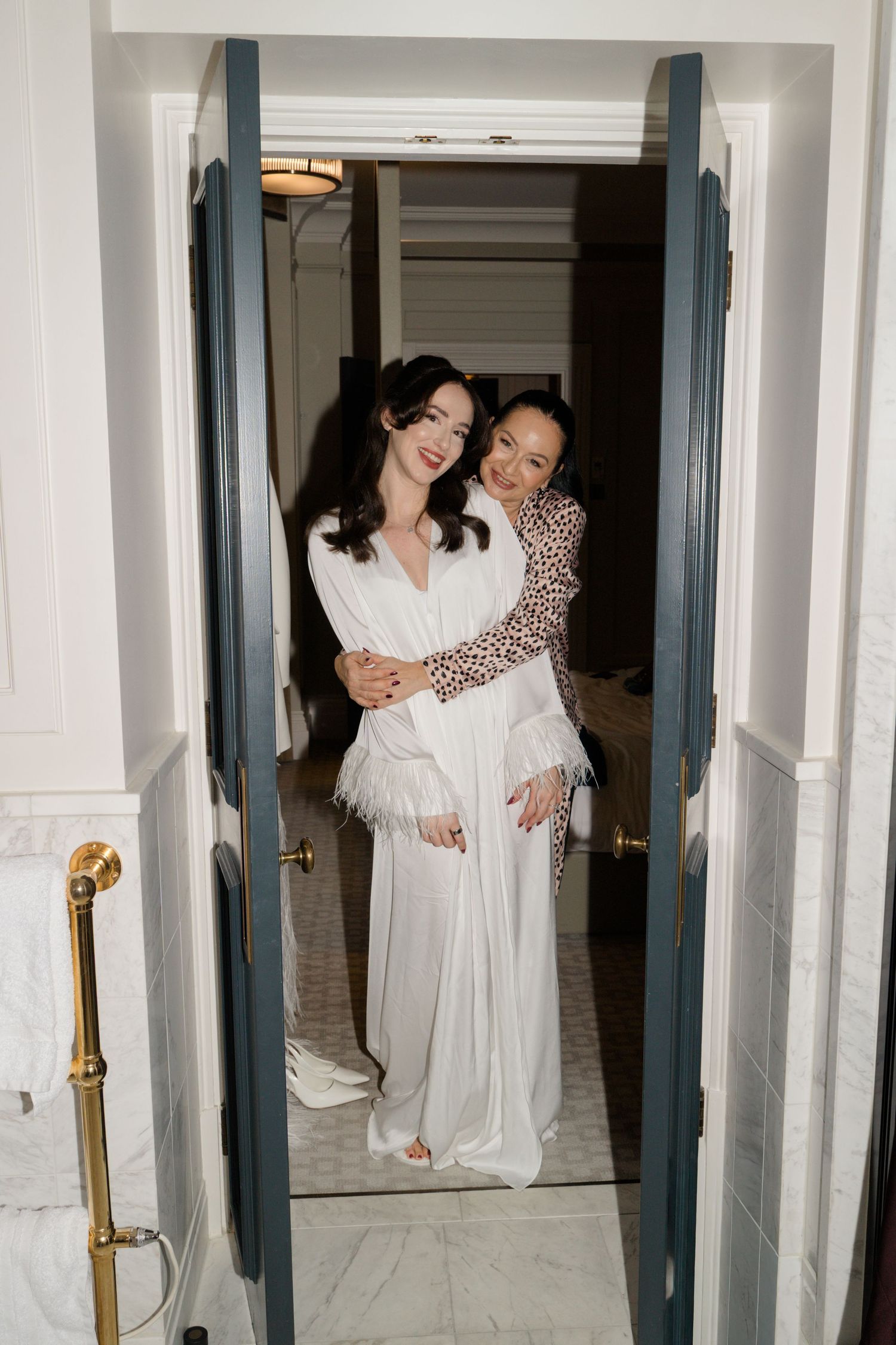 Two women in elegant white and leopard print robes embrace smiling in a doorway.