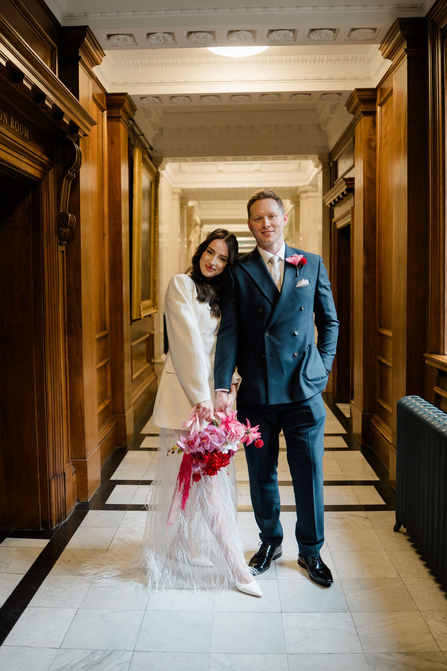 Bride and groom pose in ornate wood-paneled corridor, bride holding vibrant pink and red bouquet.
