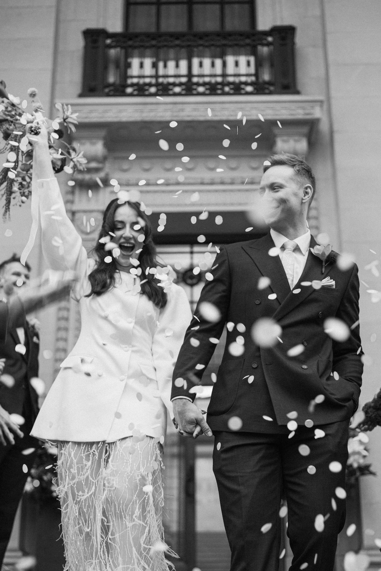 Newlyweds walk through confetti shower outside venue in joyful black and white wedding photo.