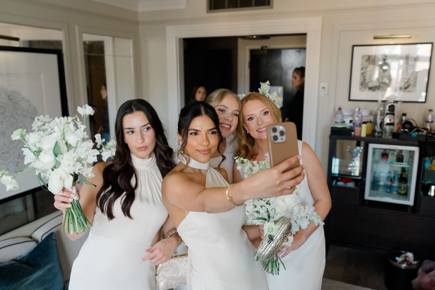 Bride and bridesmaids in white dresses taking a selfie with bouquets before wedding.