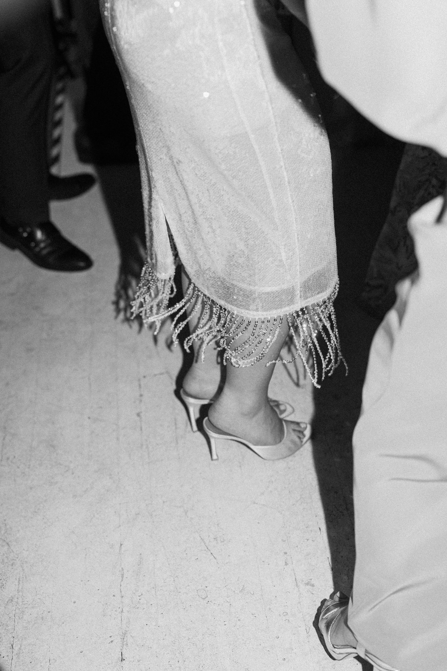 Black and white close-up of bride dancing in fringed wedding dress hem with silver heels on dance floor.