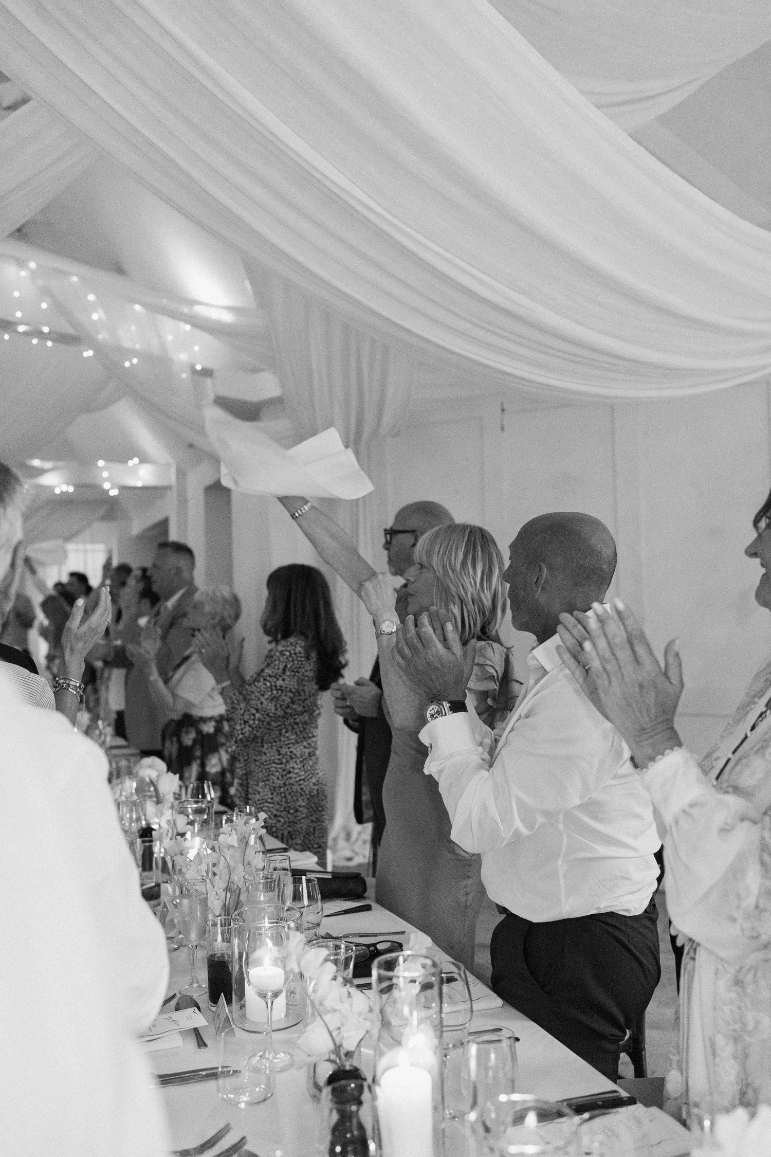 Black and white photo of wedding guests clapping and celebrating under a draped marquee reception tent.