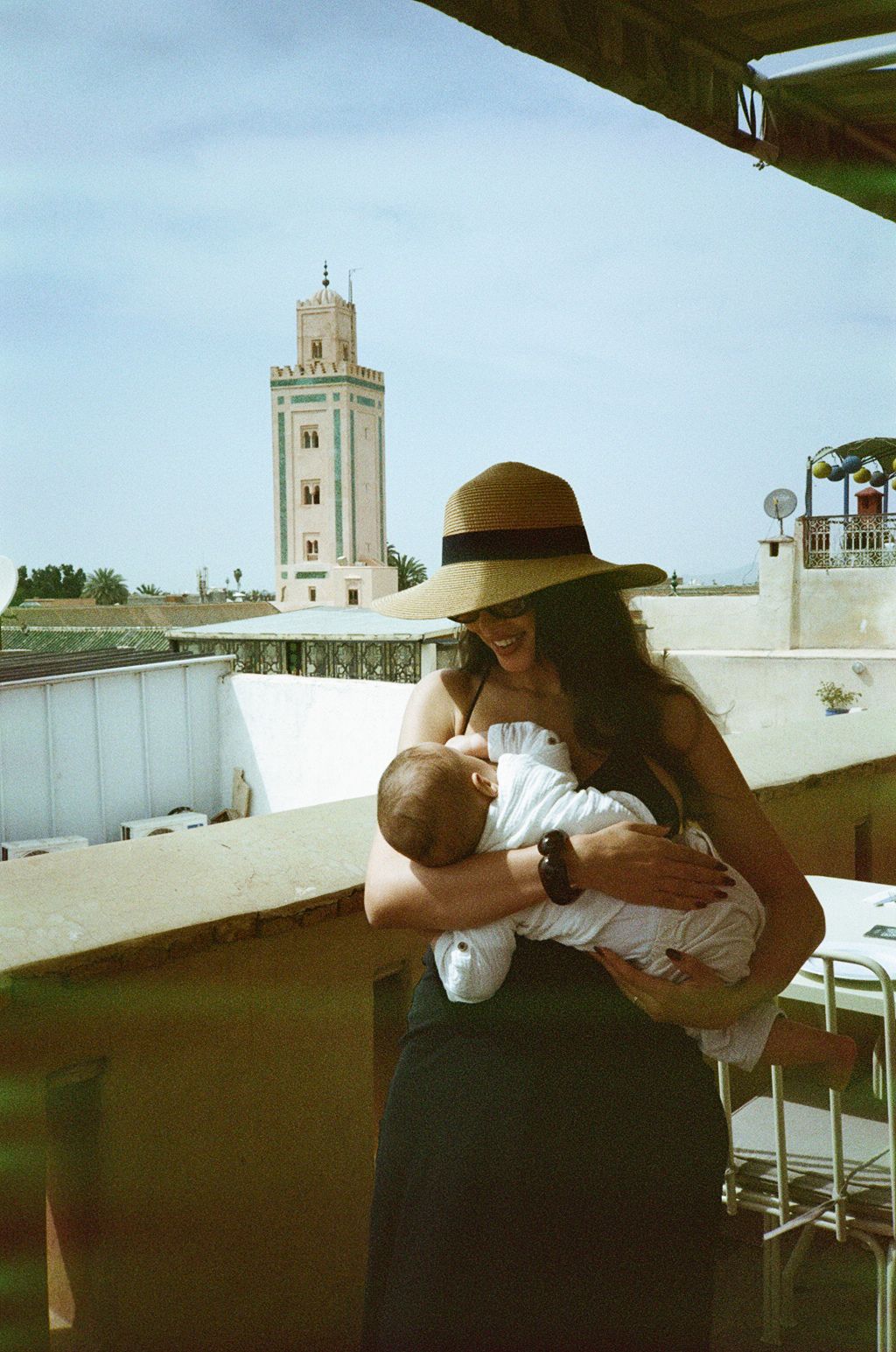 Woman in wide-brim hat breastfeeding infant on rooftop terrace with mosque minaret and blue sky in background.