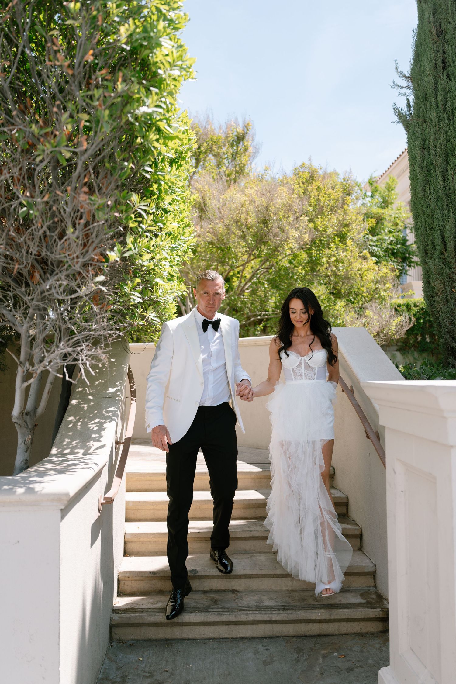 A couple descends white stone steps in wedding attire surrounded by lush greenery.