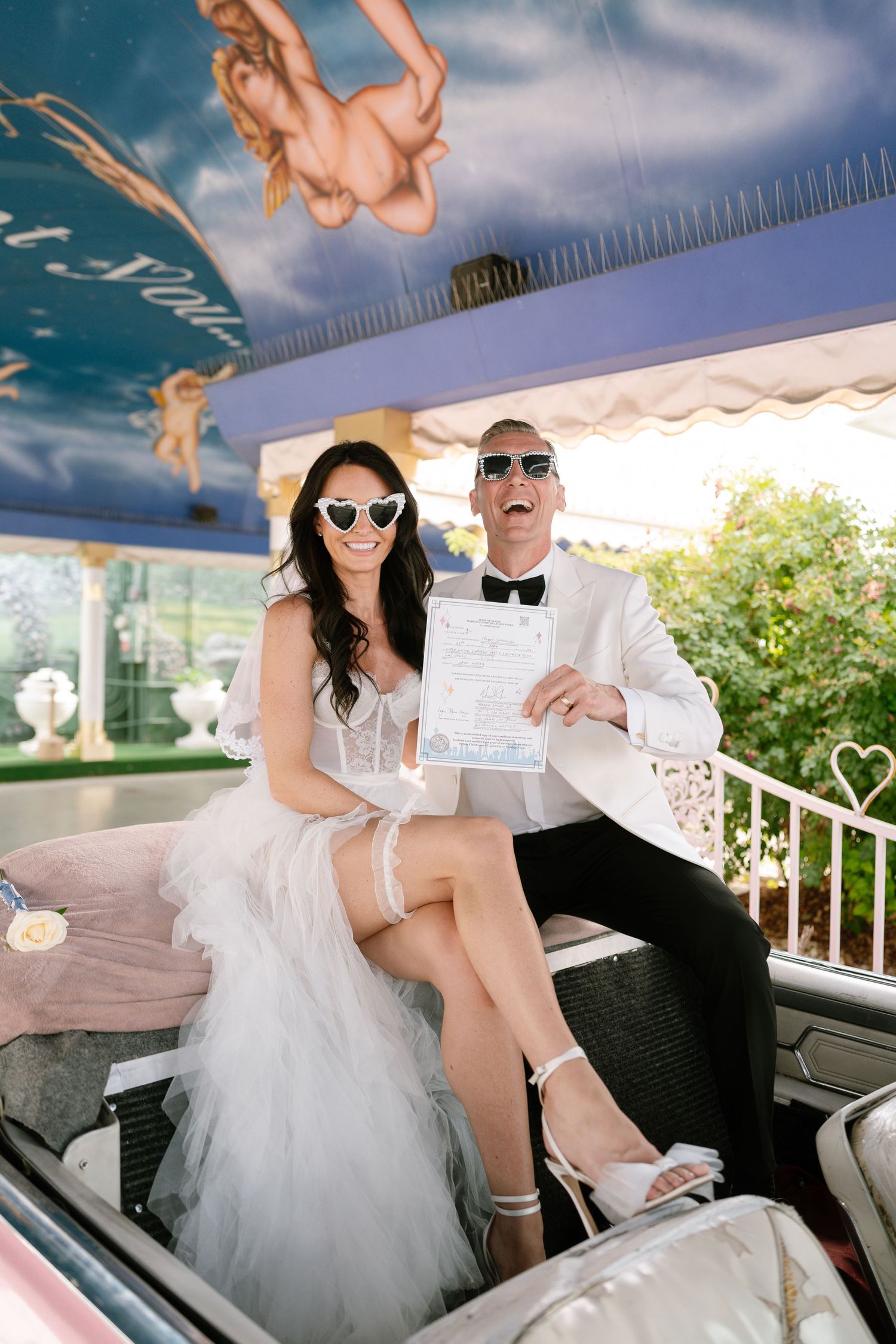 Couple wearing sunglasses poses in classic car with wedding attire.